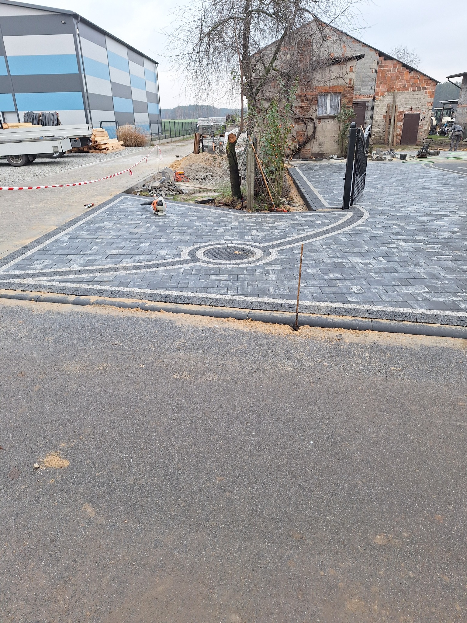 Nowo ułożona, szara kostka brukowa z dekoracyjnym wzorem koła i obramowania, prowadząca do czarnej, metalowej bramy wjazdowej; w tle budynek gospodarczy z cegły i częściowo widoczny budynek...