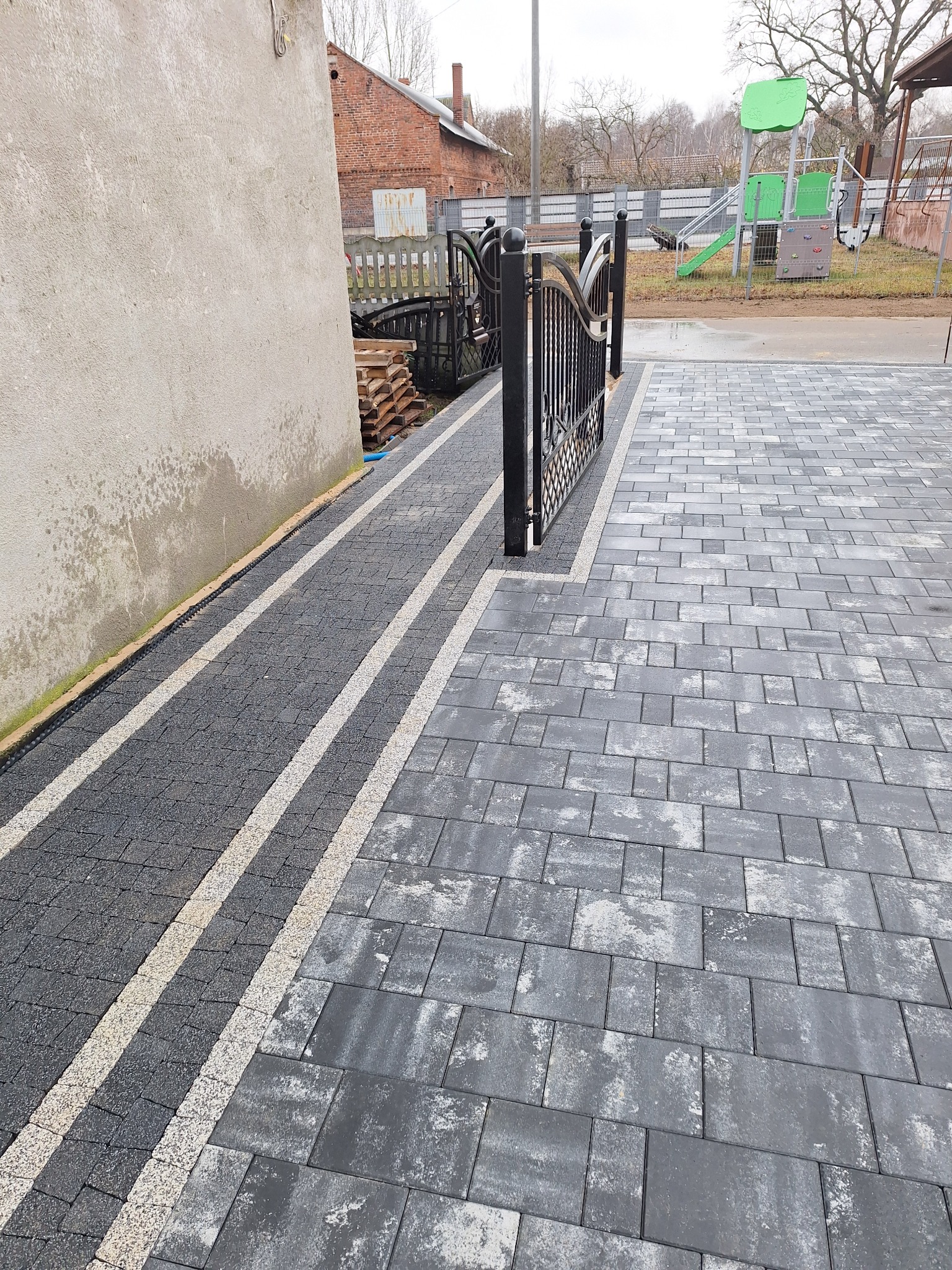 Ułożona szara kostka brukowa z jasnymi pasami oddzielającymi chodnik od podjazdu, widoczne elementy ogrodzenia i plac zabaw w tle.
