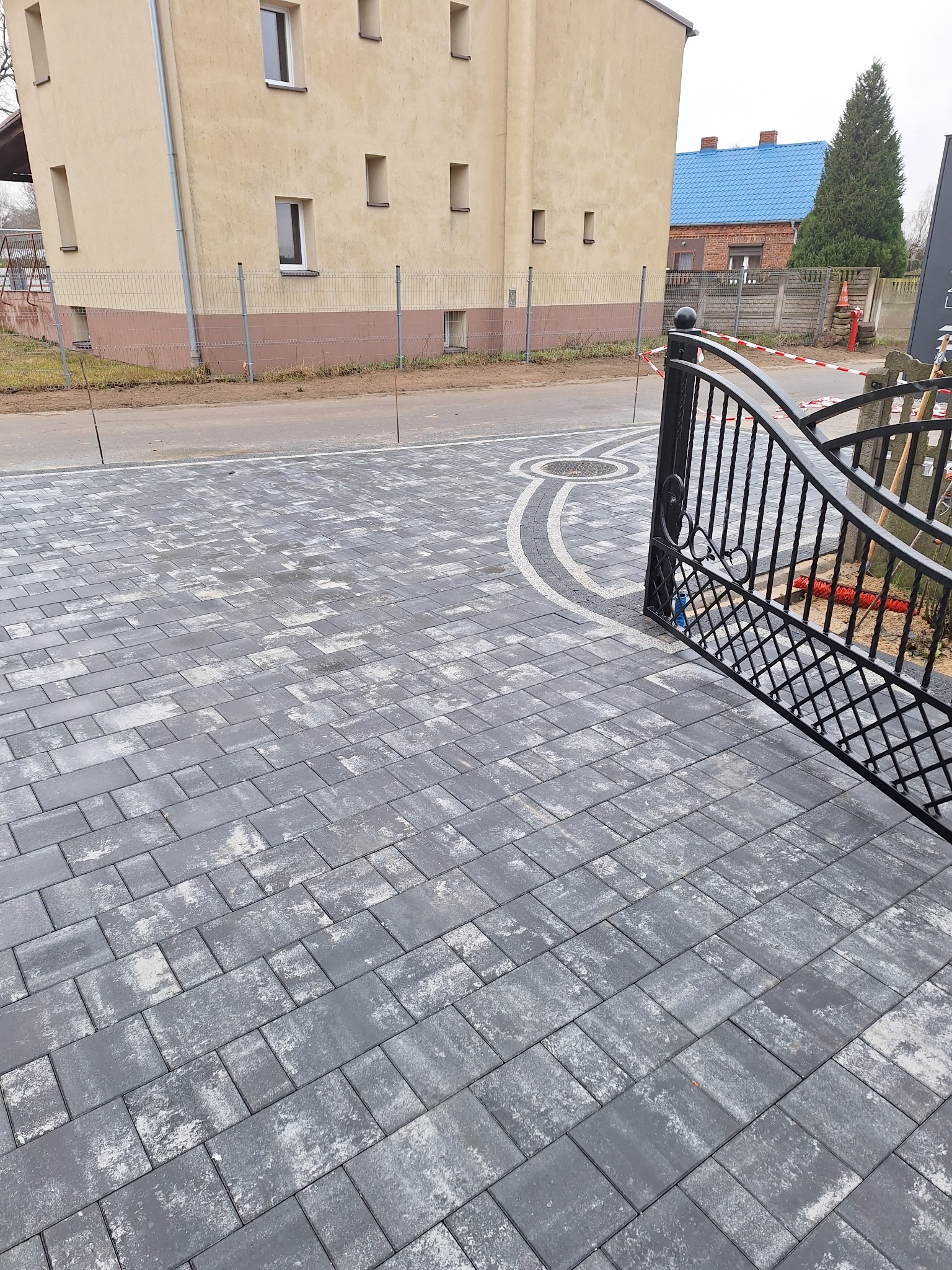 Ułożona szara kostka brukowa z dekoracyjnym okrągłym elementem w pobliżu czarnej metalowej bramy wjazdowej, w tle budynek mieszkalny.