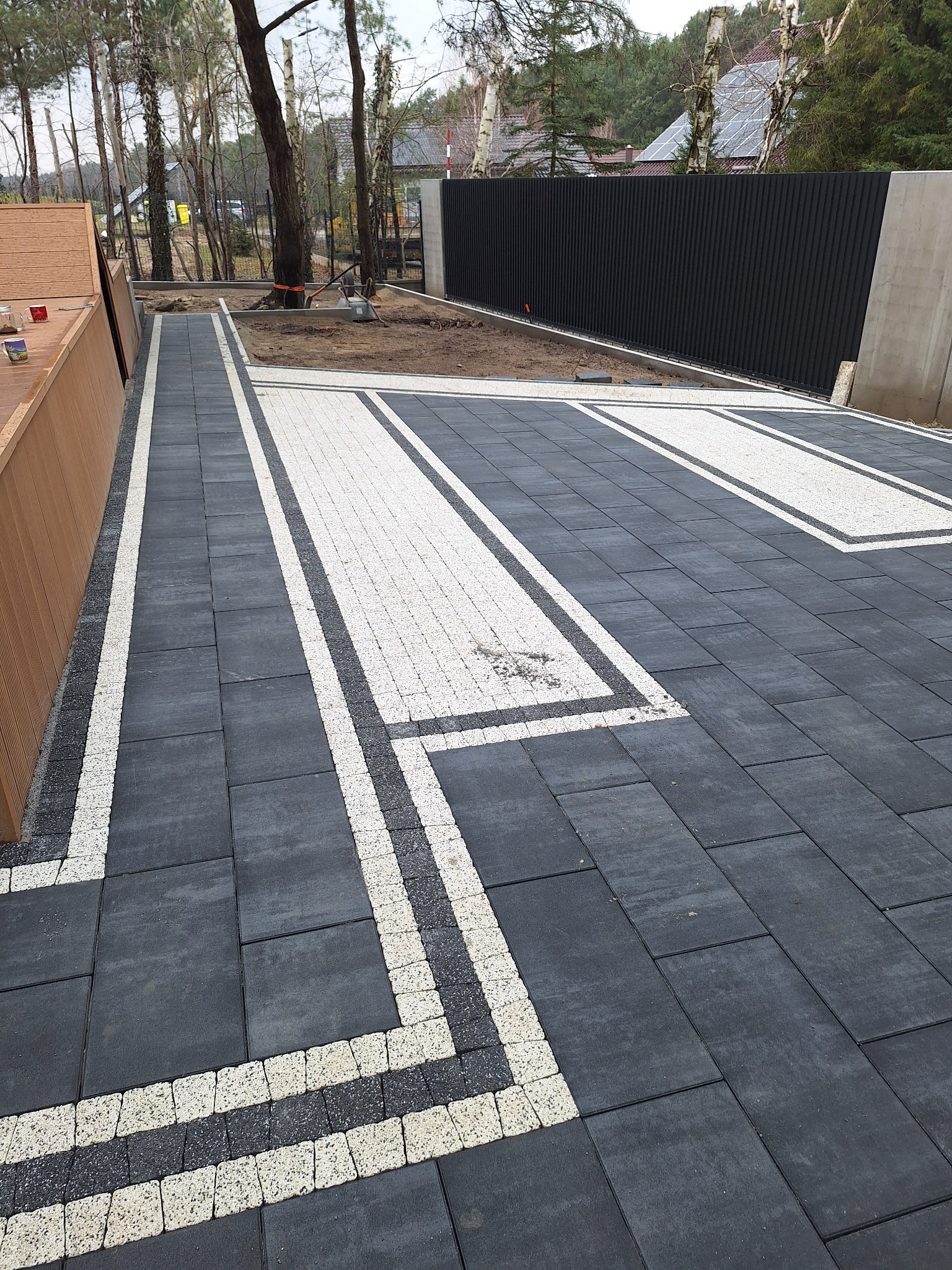 Ułożona nawierzchnia z ciemnoszarej kostki brukowej z białymi akcentami, widoczny fragment drewnianej zabudowy i czarne ogrodzenie panelowe w tle.