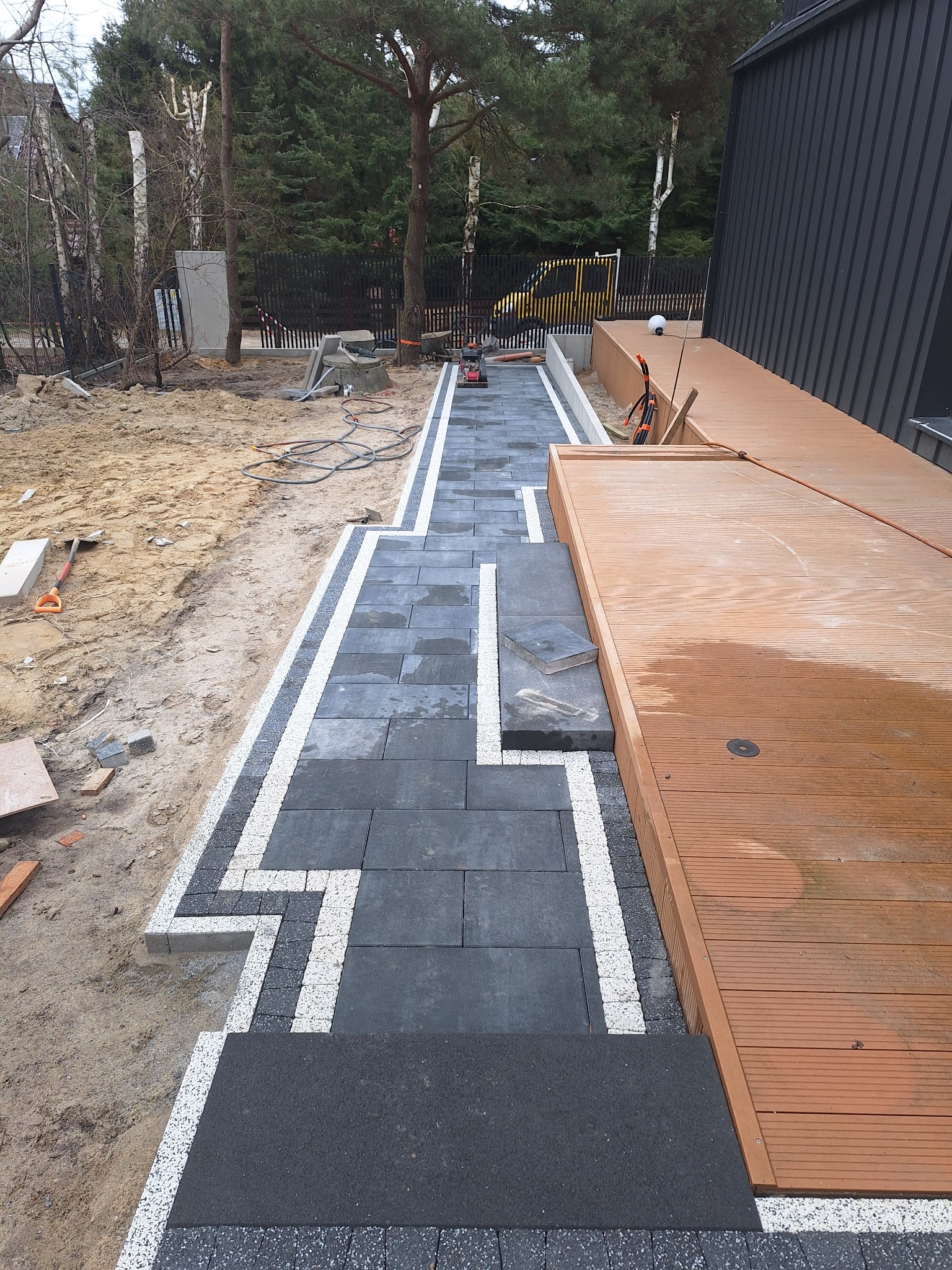 Ścieżka z ciemnoszarej kostki brukowej z białymi obramowaniami, w tle widoczna drewniana platforma i narzędzia budowlane na piaszczystym terenie.