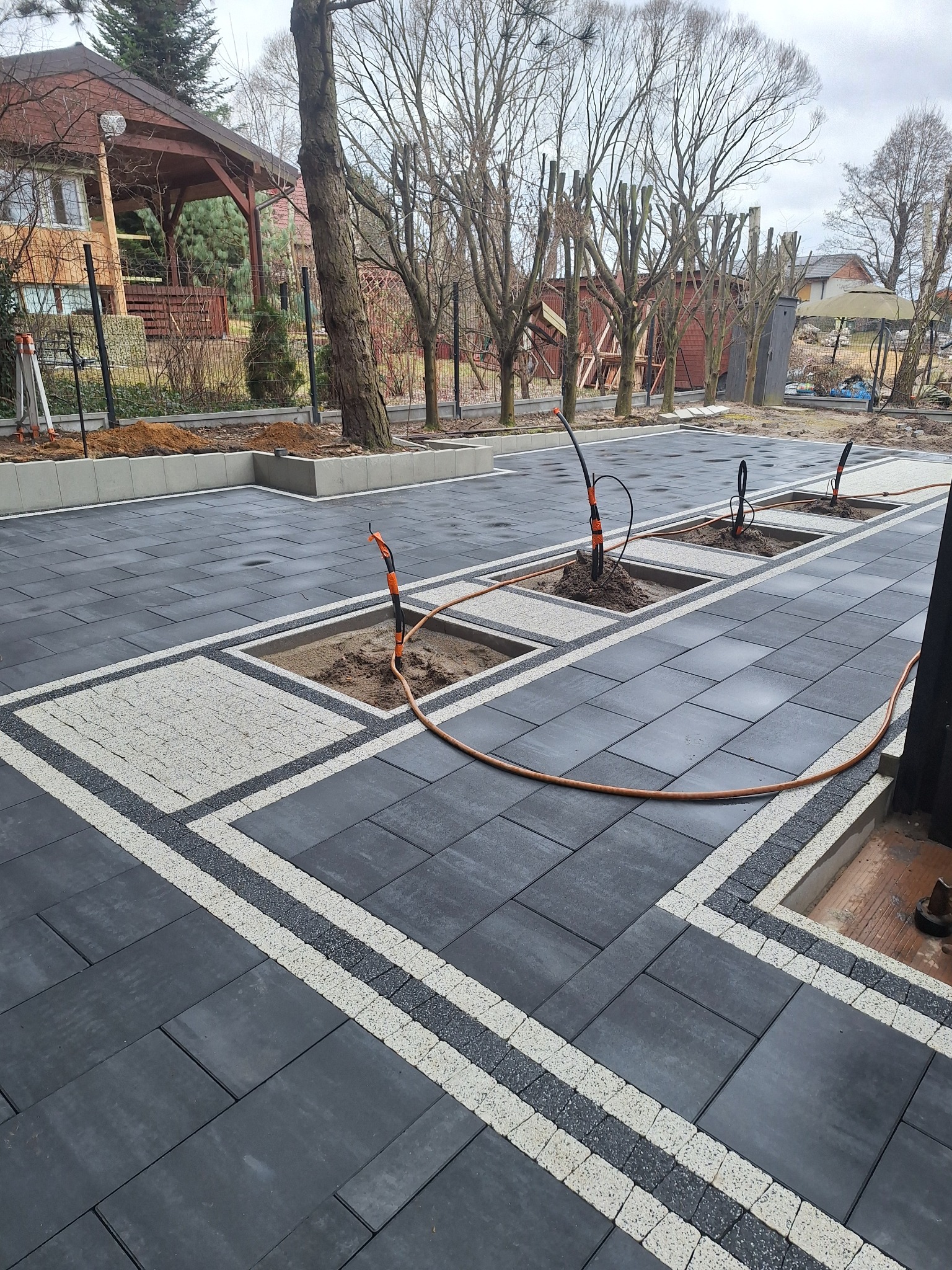 Ułożona, ciemnoszara kostka brukowa z jasnymi pasami dekoracyjnymi i miejscami na drzewa, w tle dom z drewnianą altaną i drzewa bez liści.