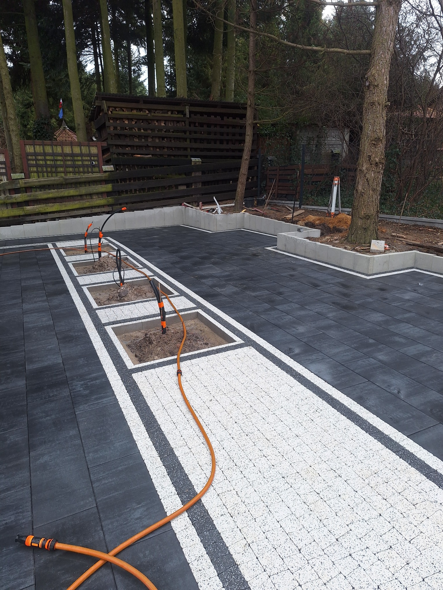 Ułożona kostka brukowa w odcieniach szarości i bieli z wydzielonymi miejscami na nasadzenia drzew, widoczny wąż ogrodowy.