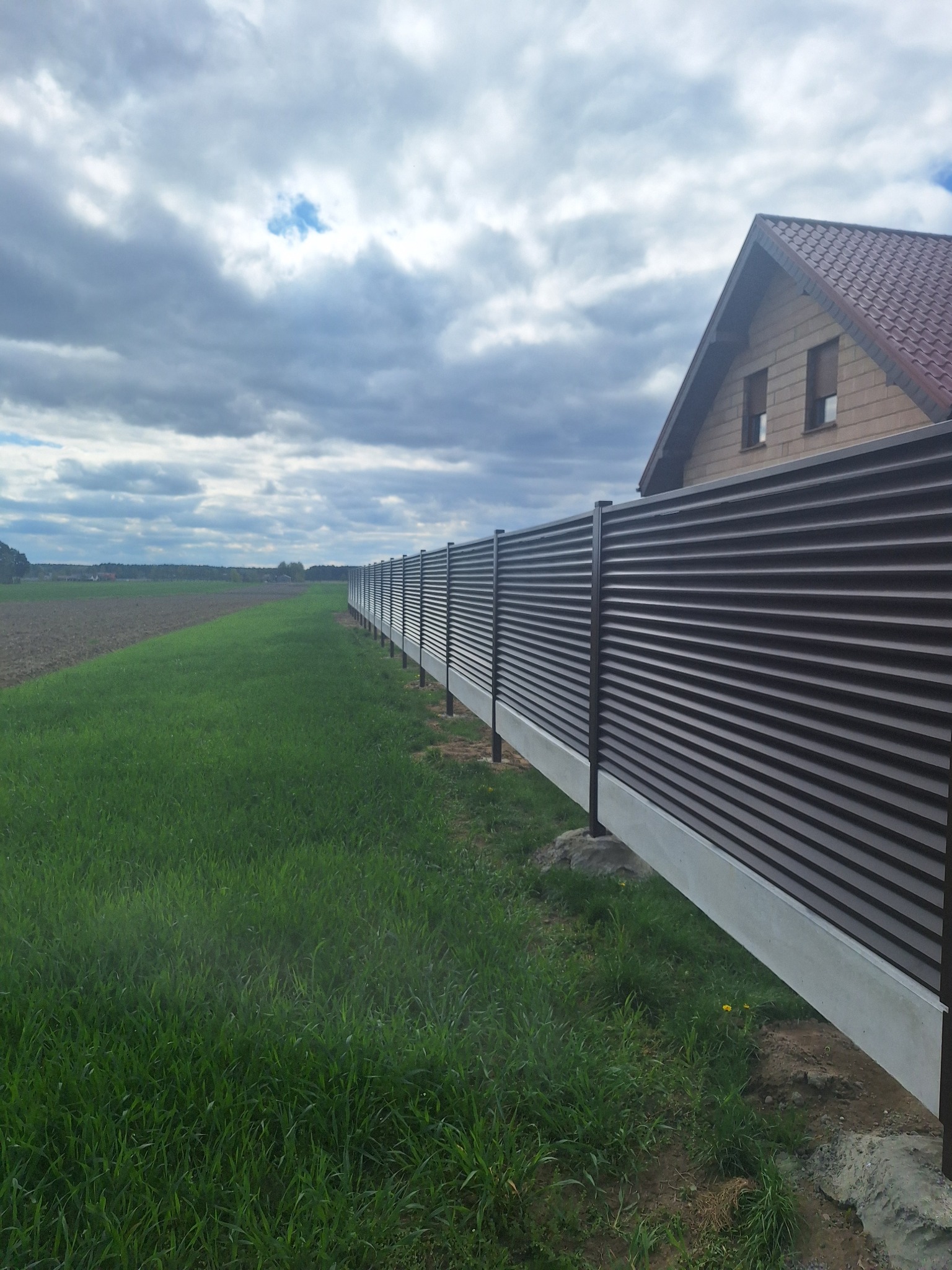Długa, brązowa, pozioma metalowa ogrodzenie panelowe z betonową podmurówką, oddzielające zielony trawnik od zaoranego pola. W tle dom z brązowym dachem i pochmurne niebo.