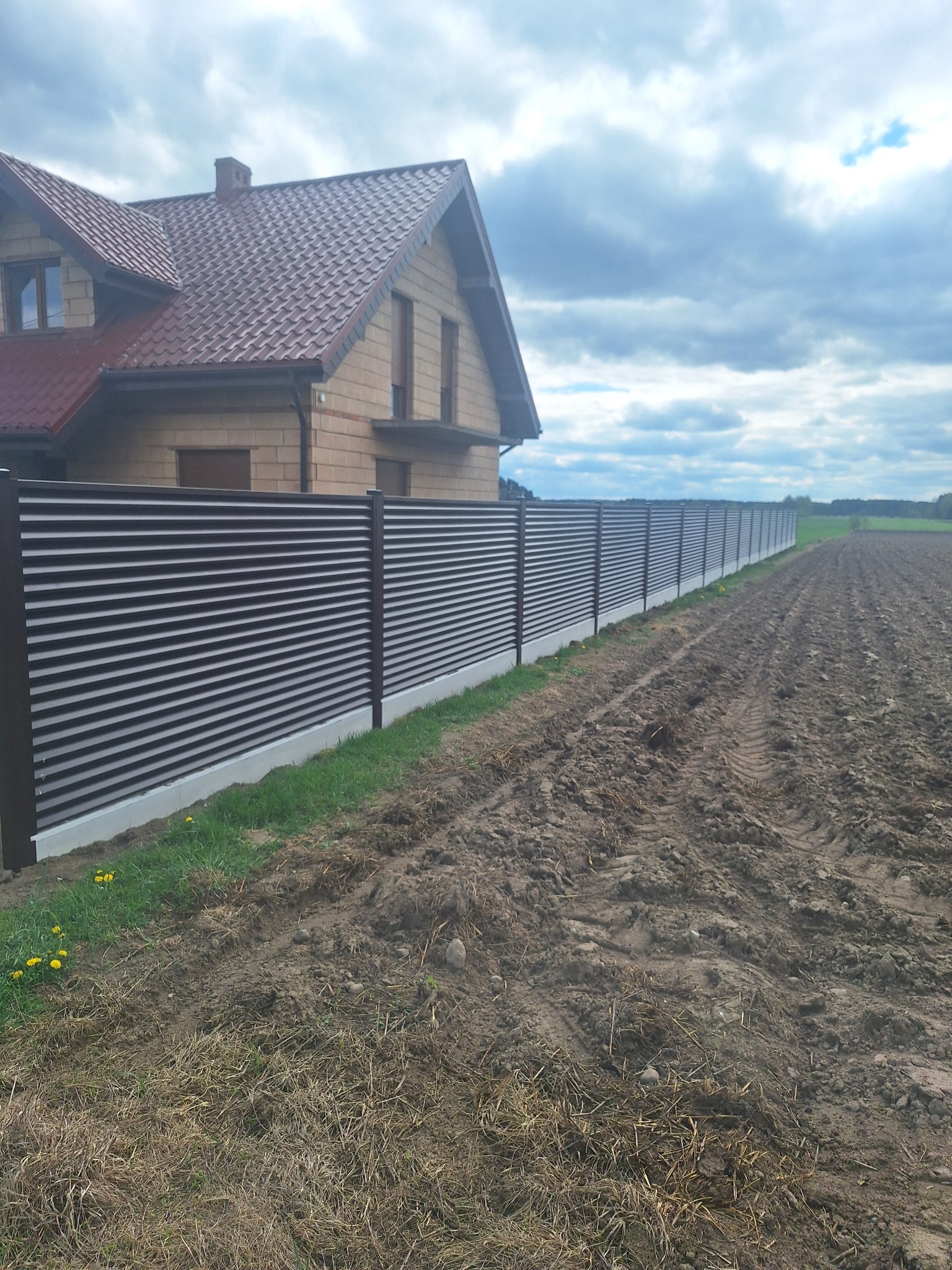 Długa, pozioma, brązowa metalowa ogrodzenie panelowe na betonowej podmurówce, oddzielające posesję z domem od pola uprawnego. Pochmurne niebo w tle.