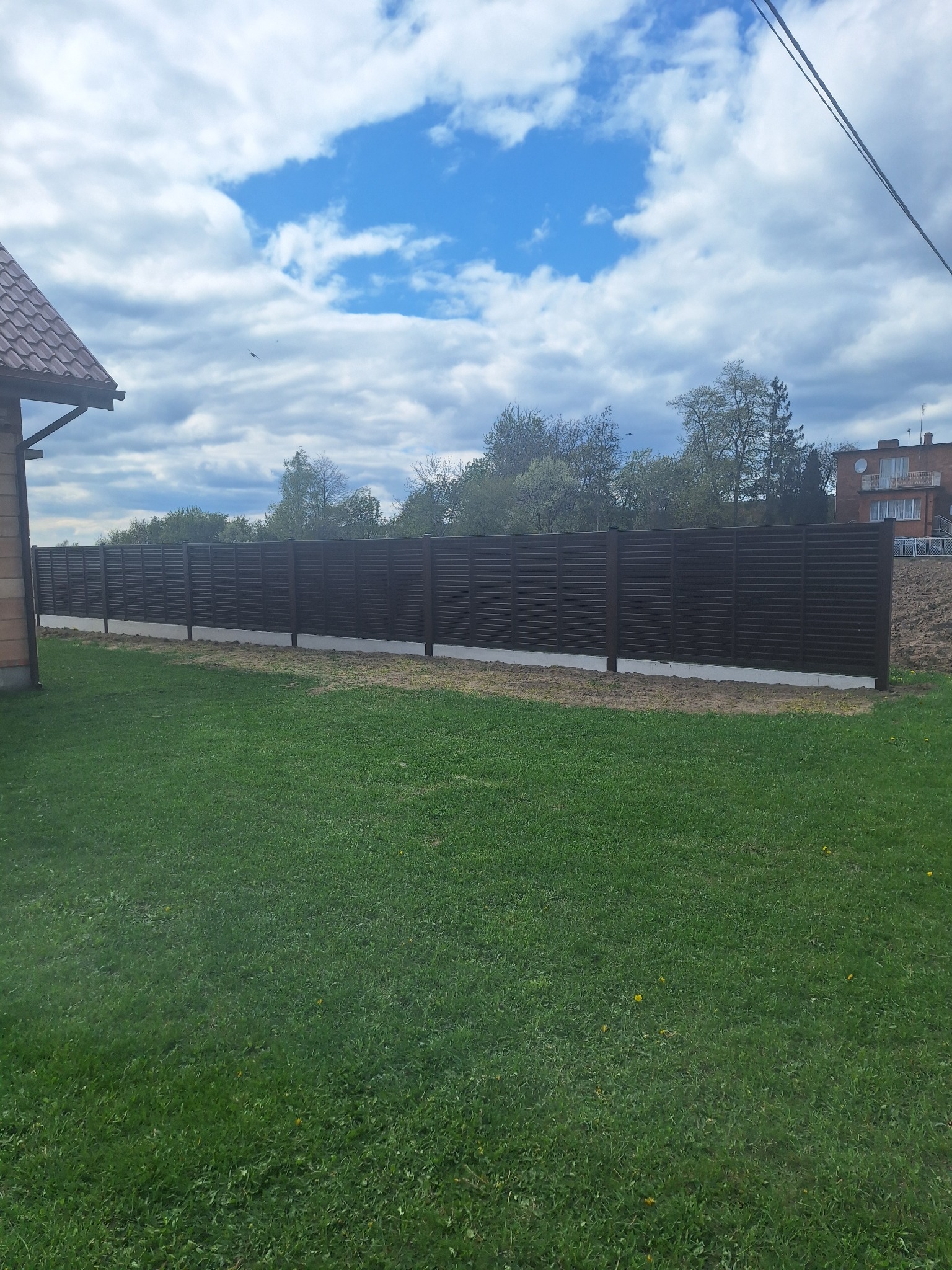 Nowoczesne, poziome, brązowe ogrodzenie panelowe na betonowej podmurówce, oddzielające posesję z zielonym trawnikiem od sąsiedniej działki.