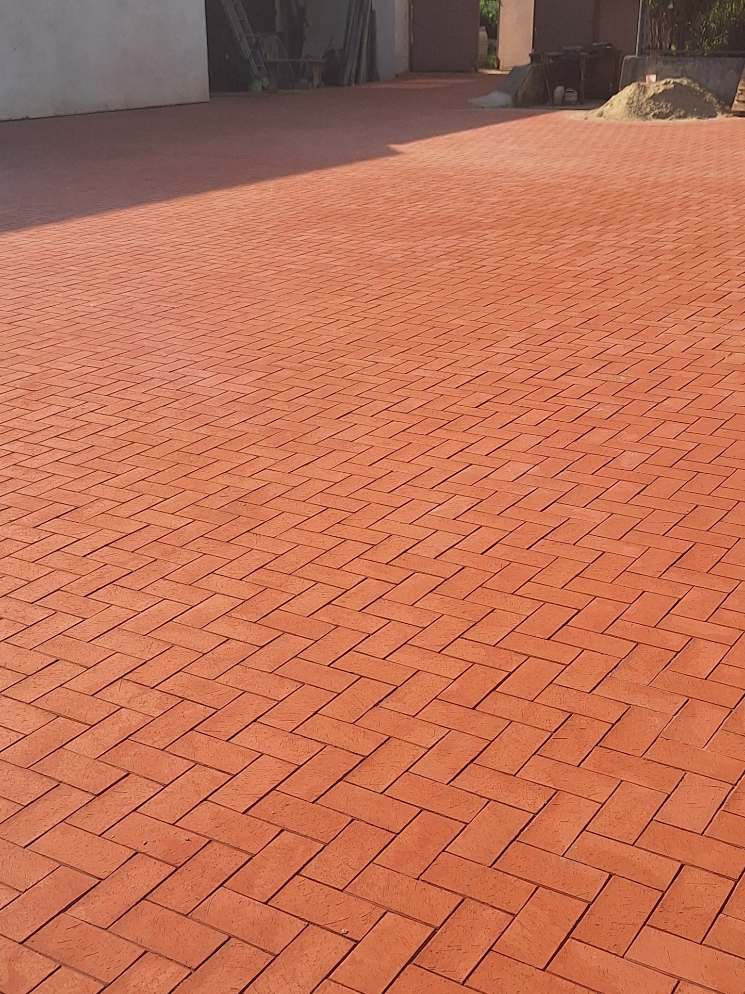Ukończona nawierzchnia z czerwonej kostki klinkierowej ułożonej we wzór jodełki, z widocznym fragmentem budynku i stertą piasku w tle.