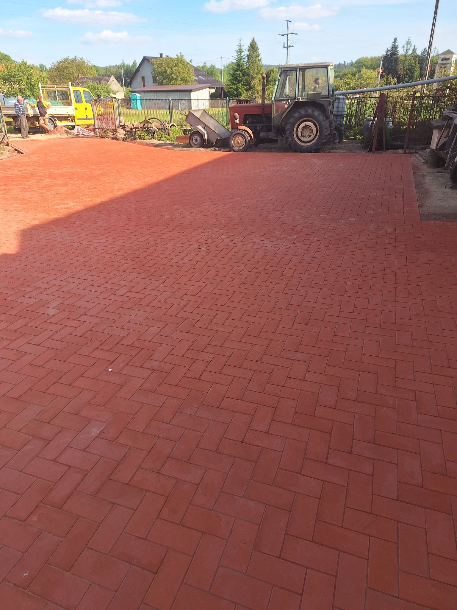Czerwona kostka brukowa ułożona na posesji, z traktorem i żółtym pojazdem w tle, widoczne budynki i drzewa, słoneczny dzień.