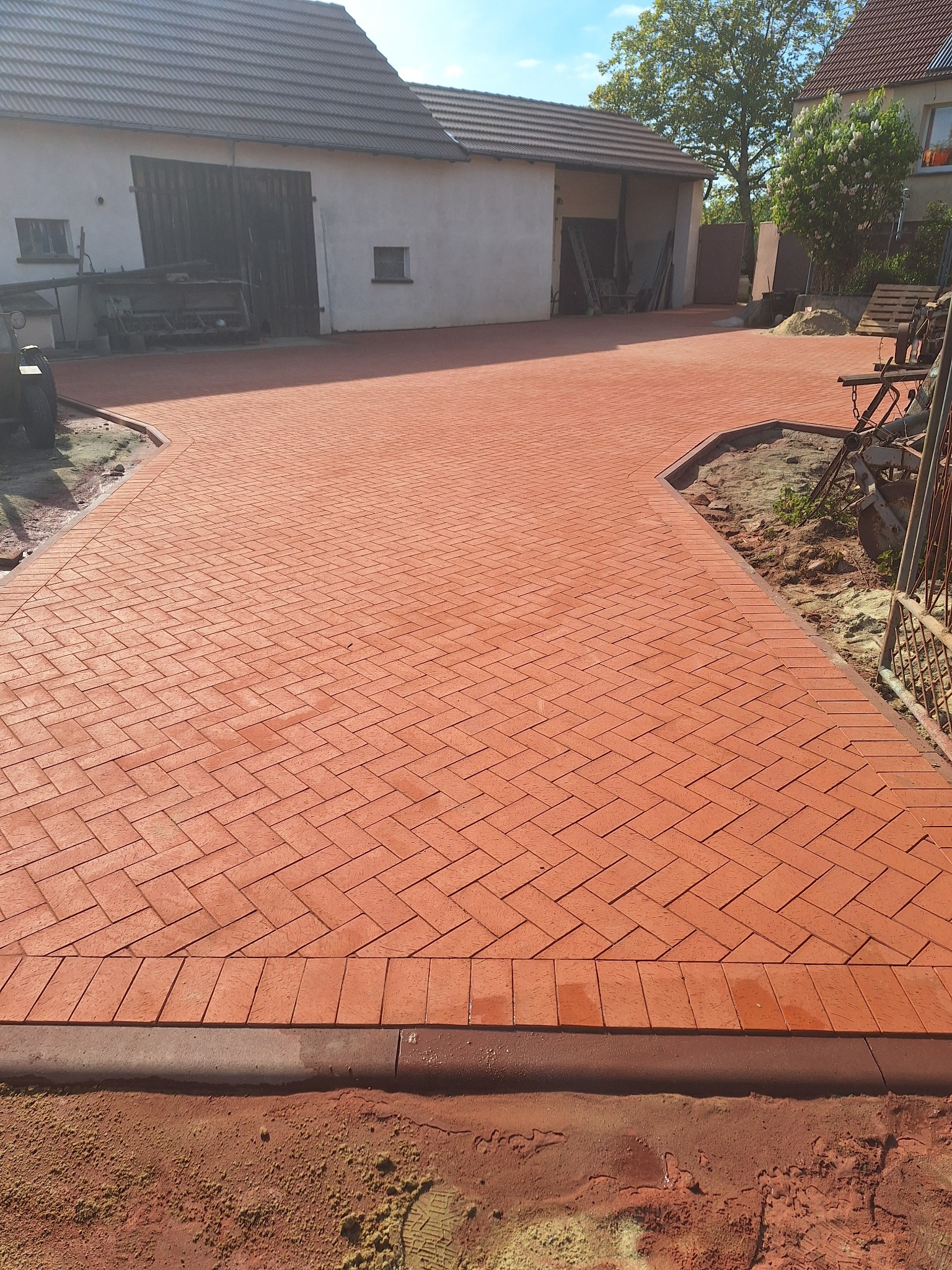 Czerwona kostka brukowa ułożona w jodełkę tworzy ścieżkę na podwórku z budynkiem gospodarczym w tle. Widoczny obrzeża i przygotowany teren pod dalsze prace.