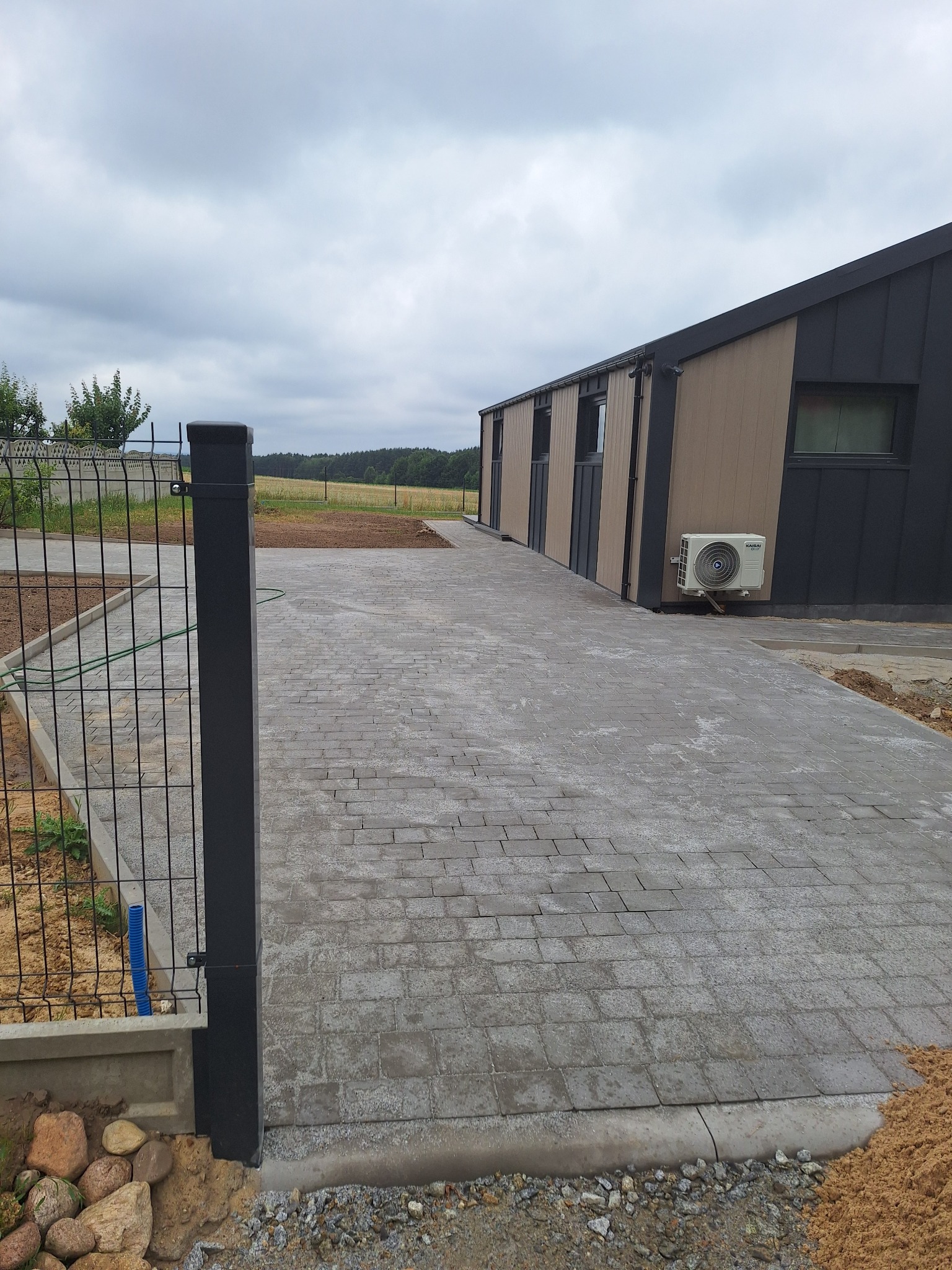 Szara kostka brukowa prowadzi do nowoczesnego budynku z klimatyzacją. W tle widoczne pole i las, a po lewej stronie ogrodzenie z siatki.