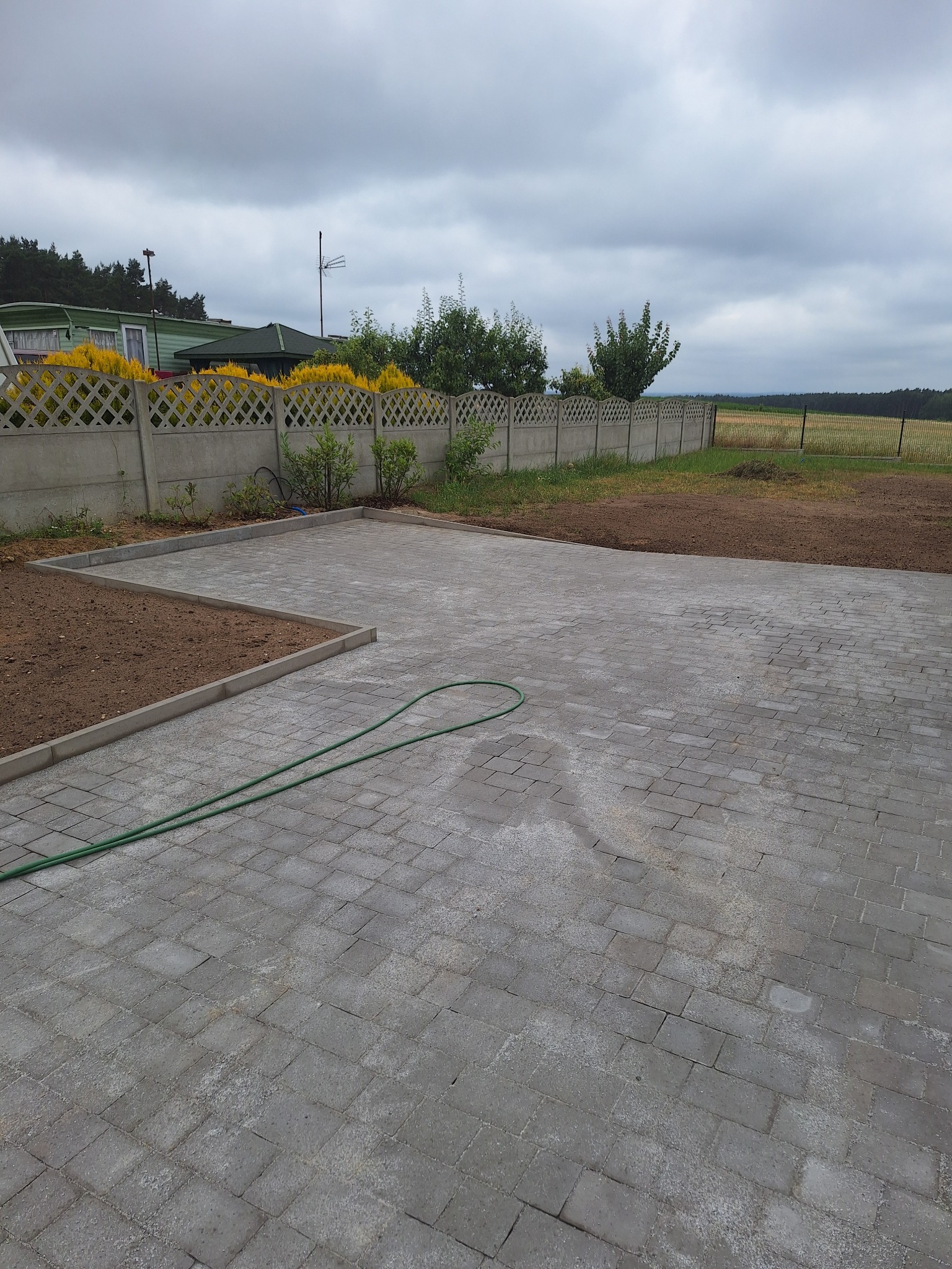 Ułożona szara kostka brukowa na podwórku, obrzeża rabatki, w tle ażurowy betonowy płot i zieleń. Wąż ogrodowy leży na kostce. Pochmurne niebo.