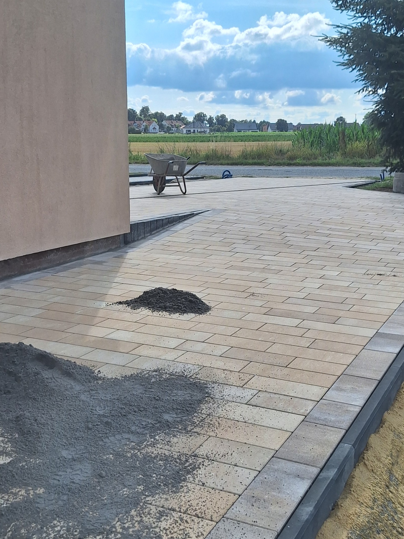 Jasna kostka brukowa w trakcie układania, widoczne pryzmy piasku i ziemi, taczka w tle, Boguszyce. Krawężniki z ciemnej kostki.