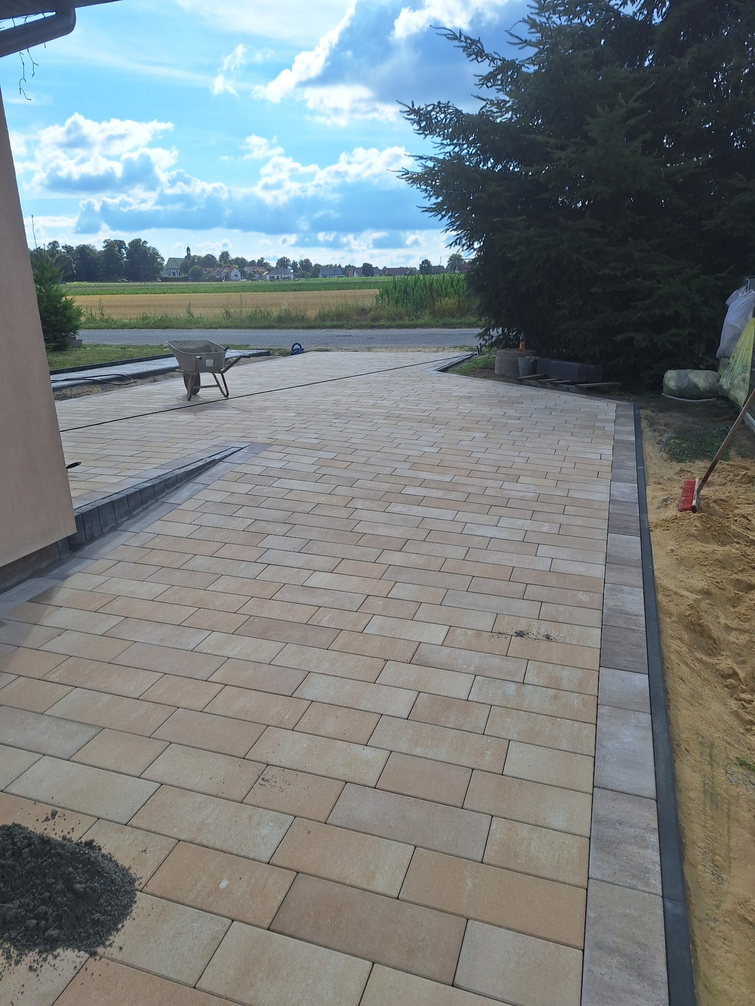 Ułożona kostka brukowa w odcieniach beżu i szarości, z widocznymi elementami wykończeniowymi. W tle krajobraz polny i błękitne niebo. Widoczny wózek i narzędzia.