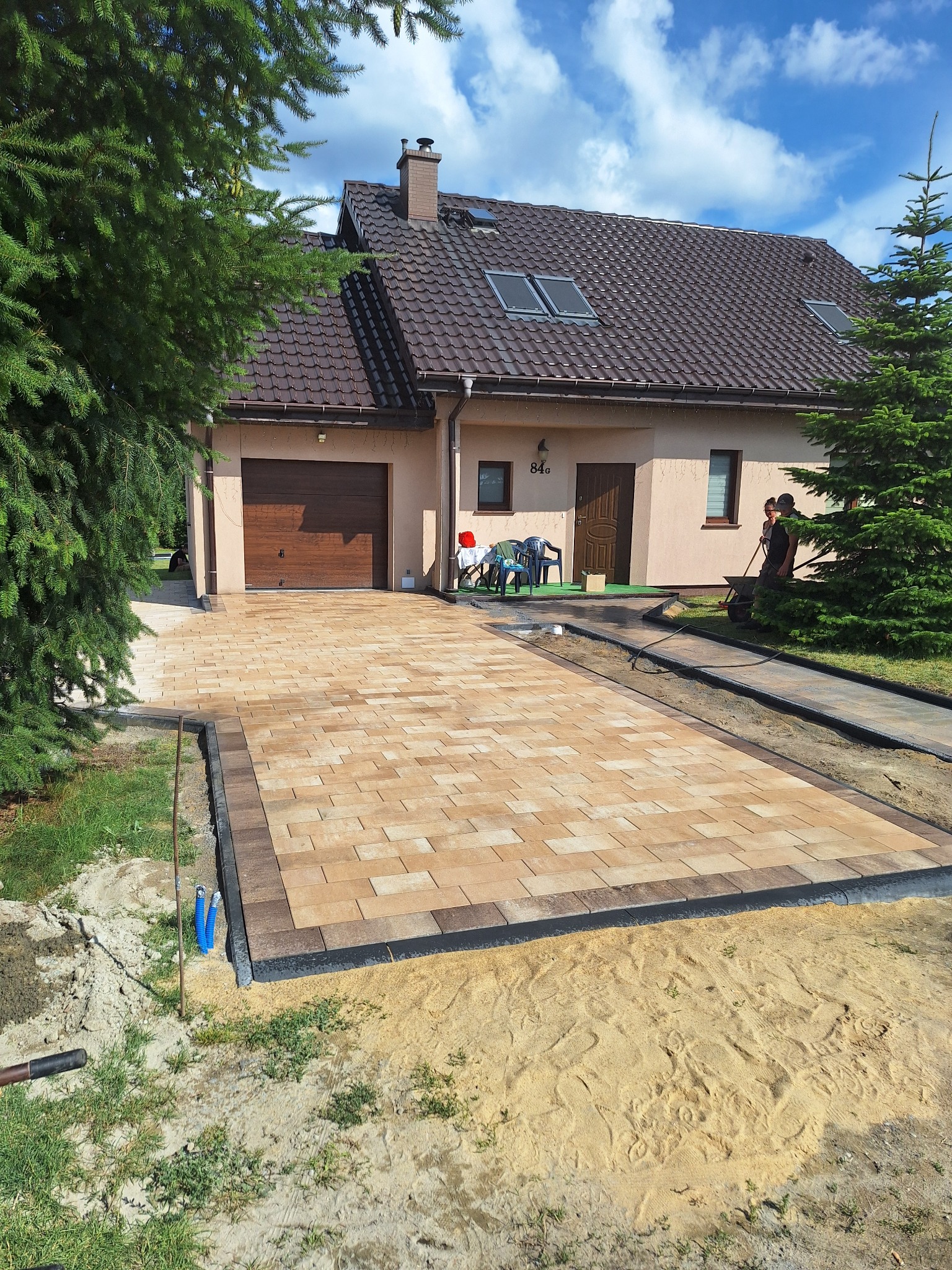 Beżowa kostka brukowa z obramowaniem przed domem z brązowym dachem. Widoczny fragment ogrodu i osoby w tle. Układanie kostki na piasku.