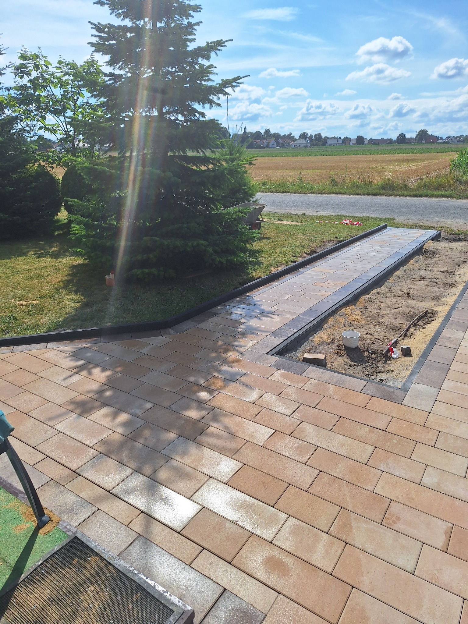 Jasnobrązowa kostka brukowa ułożona na posesji, widoczne elementy czarne obrzeża, w tle zieleń trawy i drzewa iglaste, niebo z chmurami. Częściowo ukończona praca.