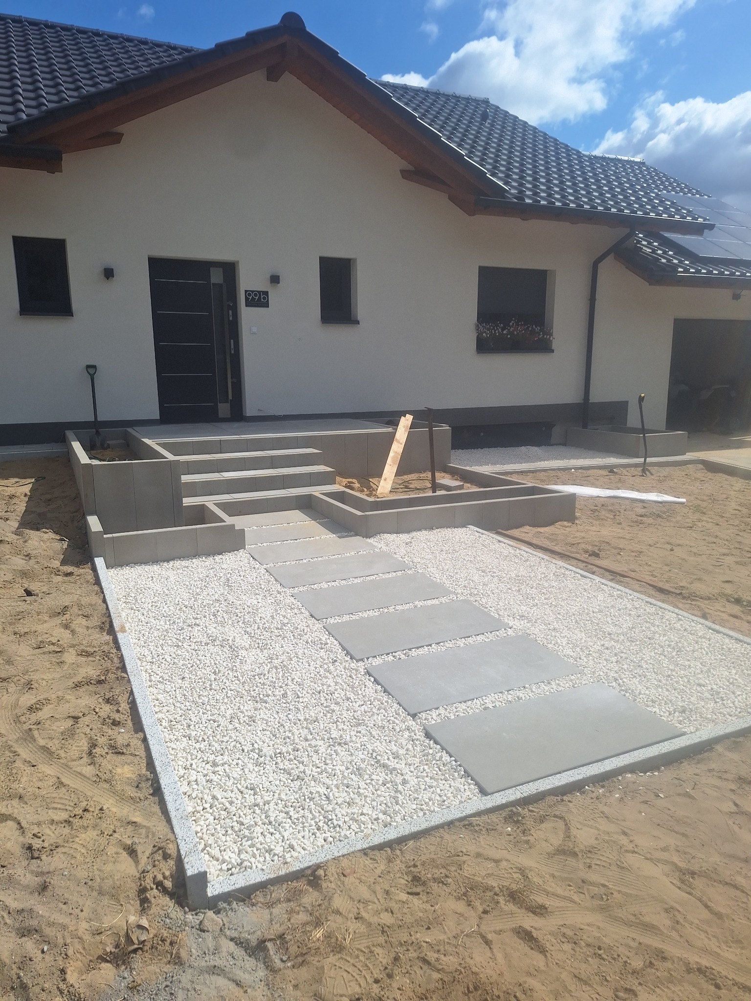 Nowoczesny ogród: ścieżka z płyt betonowych i białego grysu prowadzi do domu z schodami z kostki. Widoczne narzędzia i częściowo zrealizowane nasadzenia.