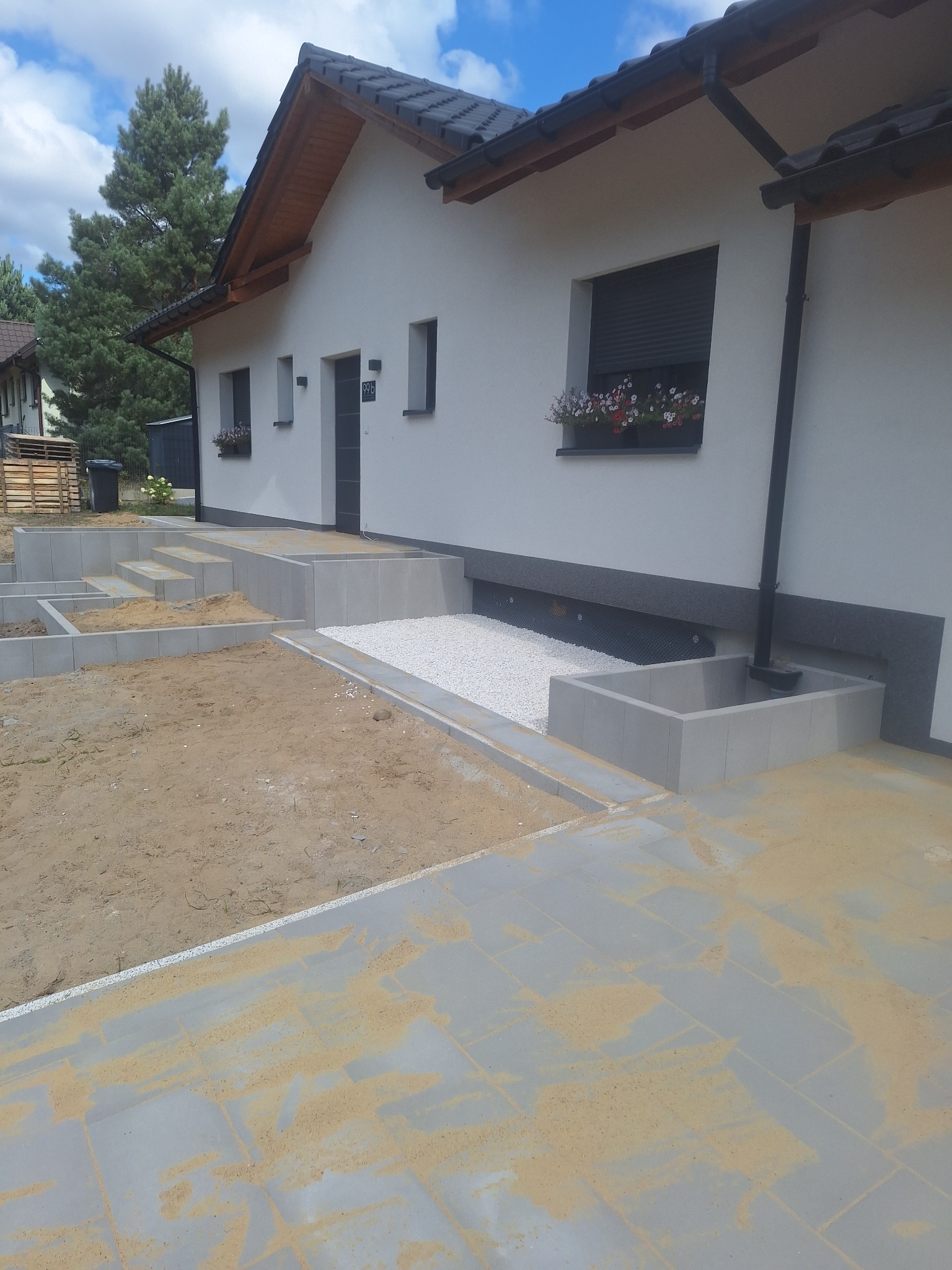 Nowoczesny dom z szarą kostką brukową, schody z betonowych bloczków i rabata z białym żwirem. Prace brukarskie w toku, widoczny piasek na kostce. Słoneczny dzień.