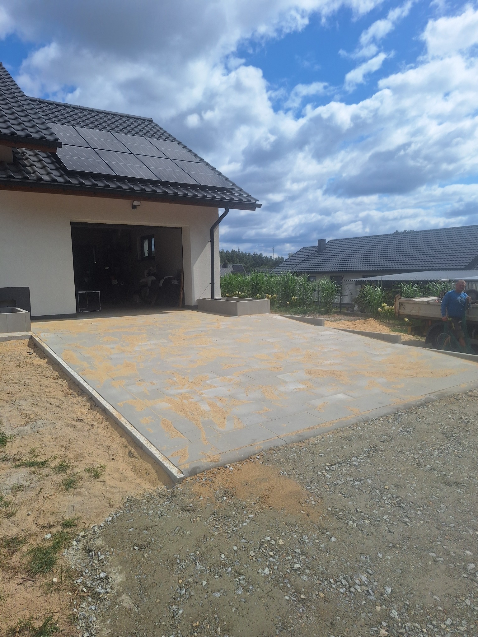 Jasnoszara kostka brukowa ułożona przed garażem domu z panelami słonecznymi na dachu, posypana piaskiem. W tle widoczny mężczyzna i samochód dostawczy.