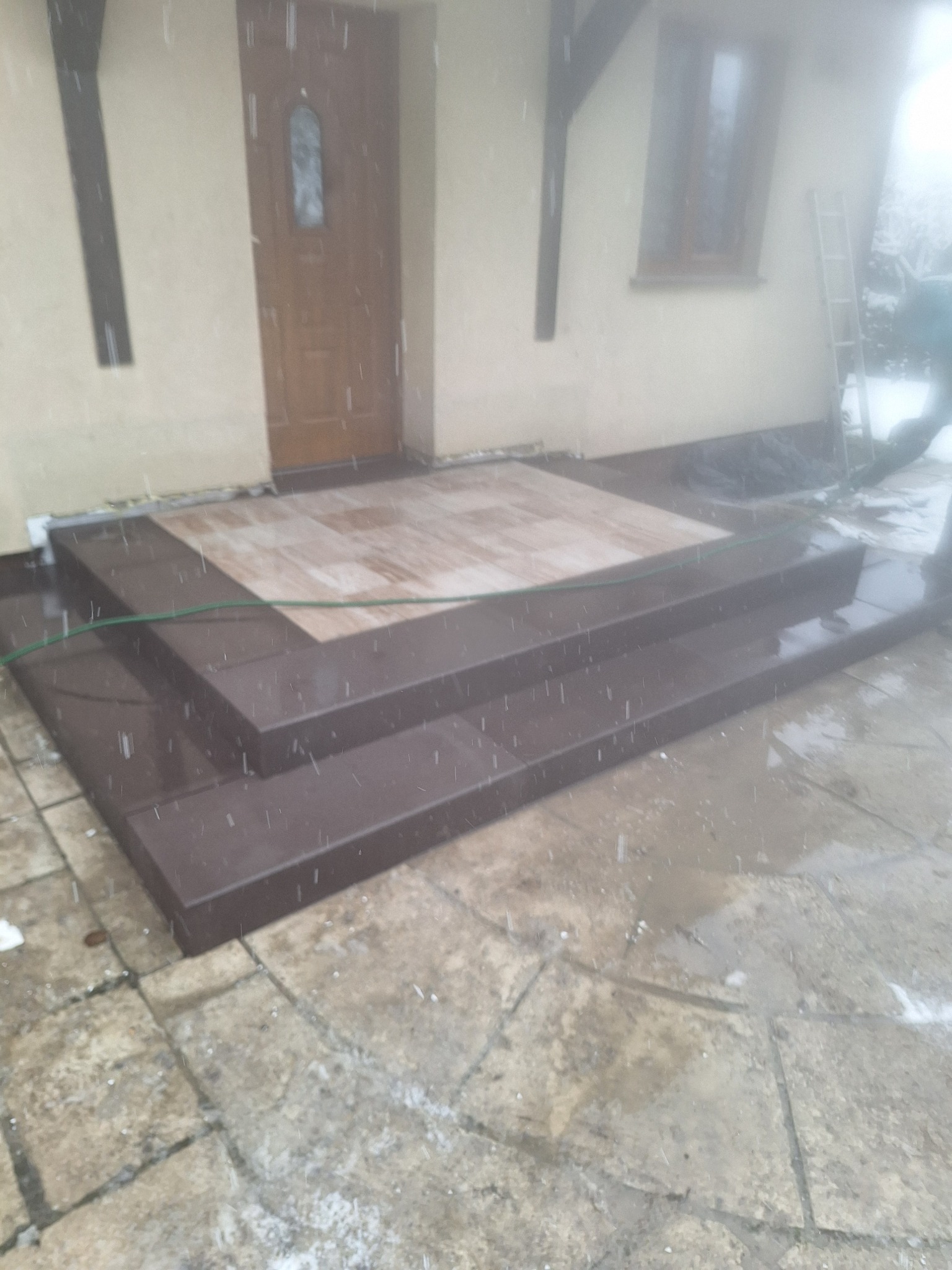 Wykończone schody wejściowe z brązowych płytek, prowadzące do drzwi domu. Kostka brukowa na podłożu. Widoczne opady deszczu lub śniegu.