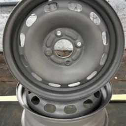 Dwie stalowe felgi samochodowe, jedna na drugiej, po obróbce, z widoczną szarą, matową powierzchnią i otworami wentylacyjnymi, na tle ściany z cegieł i fluorescencyjnej lampy.