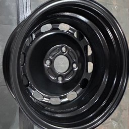 AMP - Pomalowana na czarno felga samochodowa, zawieszona na metalowej konstrukcji w warsztacie.