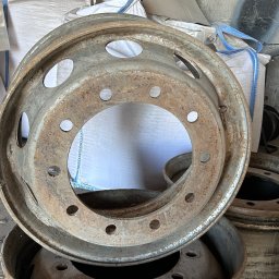 AMP - Stare, zardzewiałe felgi samochodowe z licznymi otworami, ułożone jedna na drugiej, z workami w tle, przed piaskowaniem.