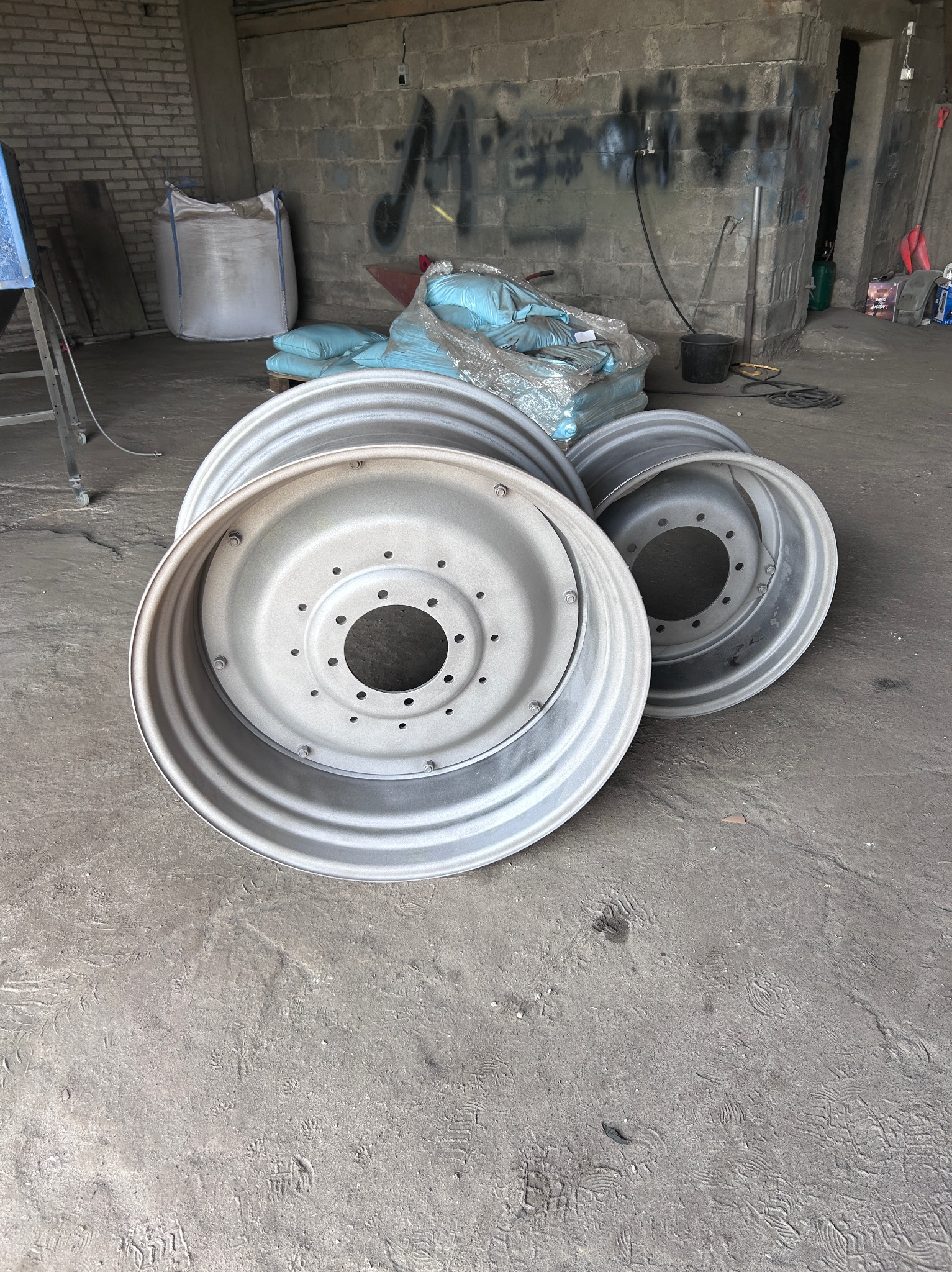 Trzy metalowe felgi po obróbce, leżące na betonowej podłodze w warsztacie, z workami w tle. Widoczne ślady użytkowania na podłodze i graffiti na ścianie.