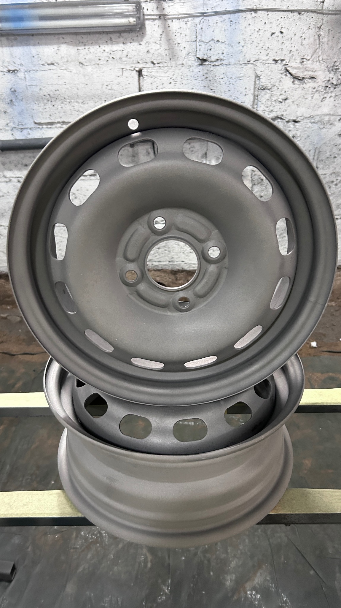 Dwie stalowe felgi samochodowe, jedna na drugiej, po obróbce, z widoczną szarą, matową powierzchnią i otworami wentylacyjnymi, na tle ściany z cegieł i fluorescencyjnej lampy.