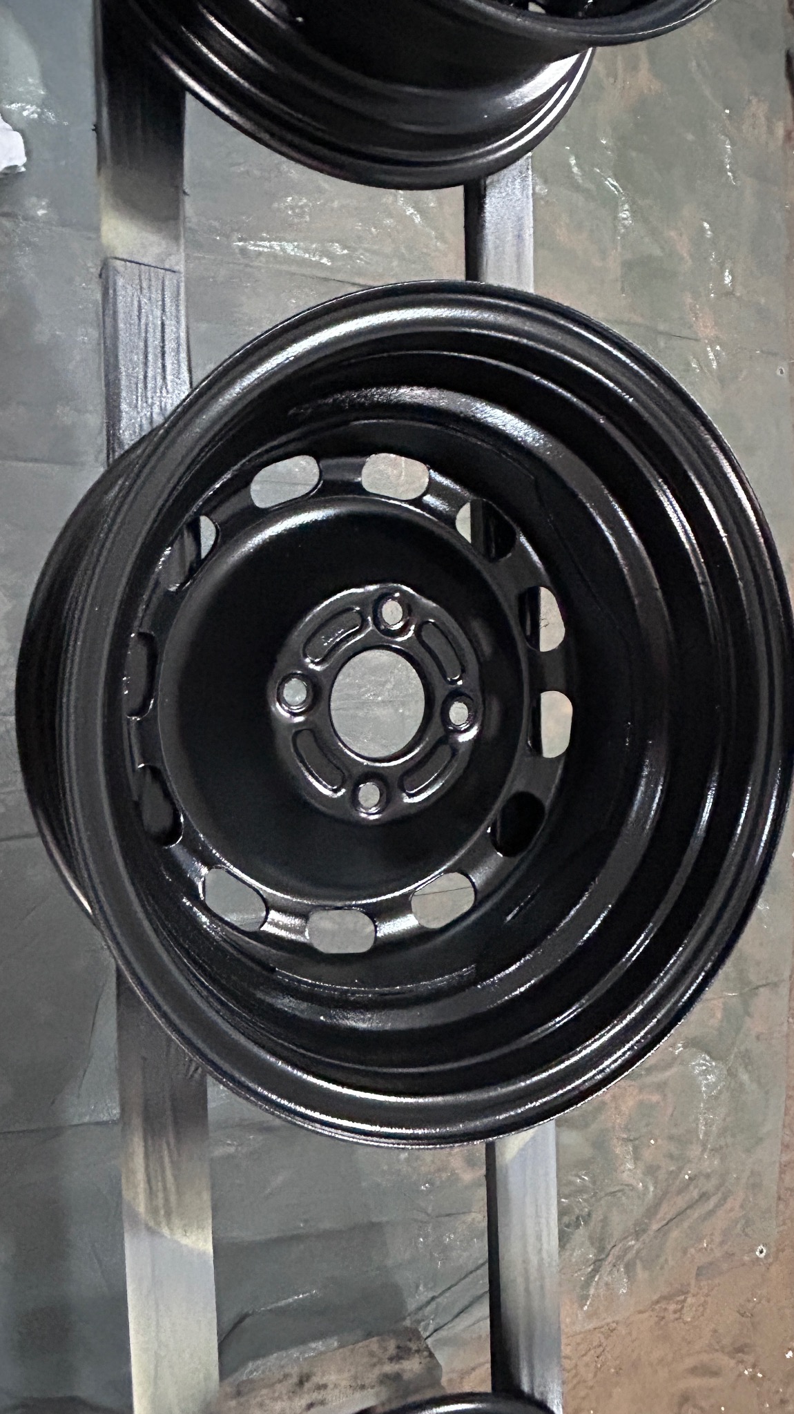 Pomalowana na czarno felga samochodowa, zawieszona na metalowej konstrukcji w warsztacie.