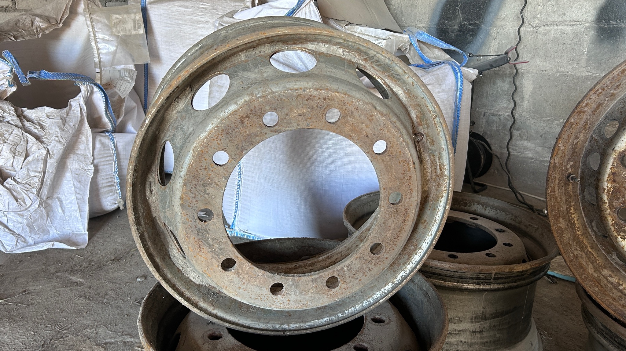 Stare, zardzewiałe felgi samochodowe z licznymi otworami, ułożone jedna na drugiej, z workami w tle, przed piaskowaniem.