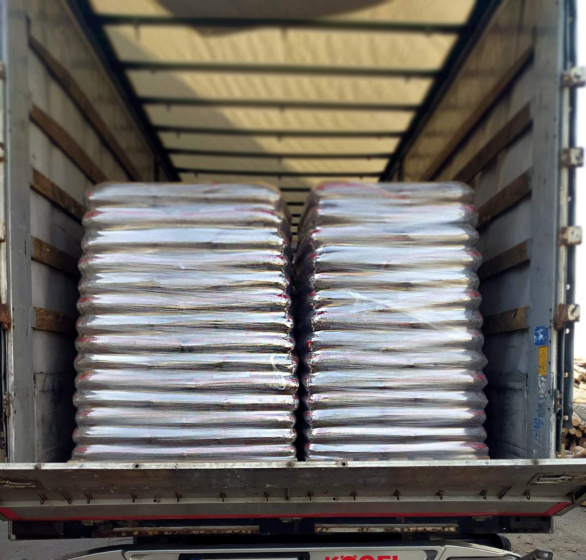 Dwa rzędy palet z workami opału, zabezpieczonych folią, widoczne w przestrzeni ładunkowej ciężarówki z otwartymi drzwiami; w tle, na zewnątrz, stos drewna.