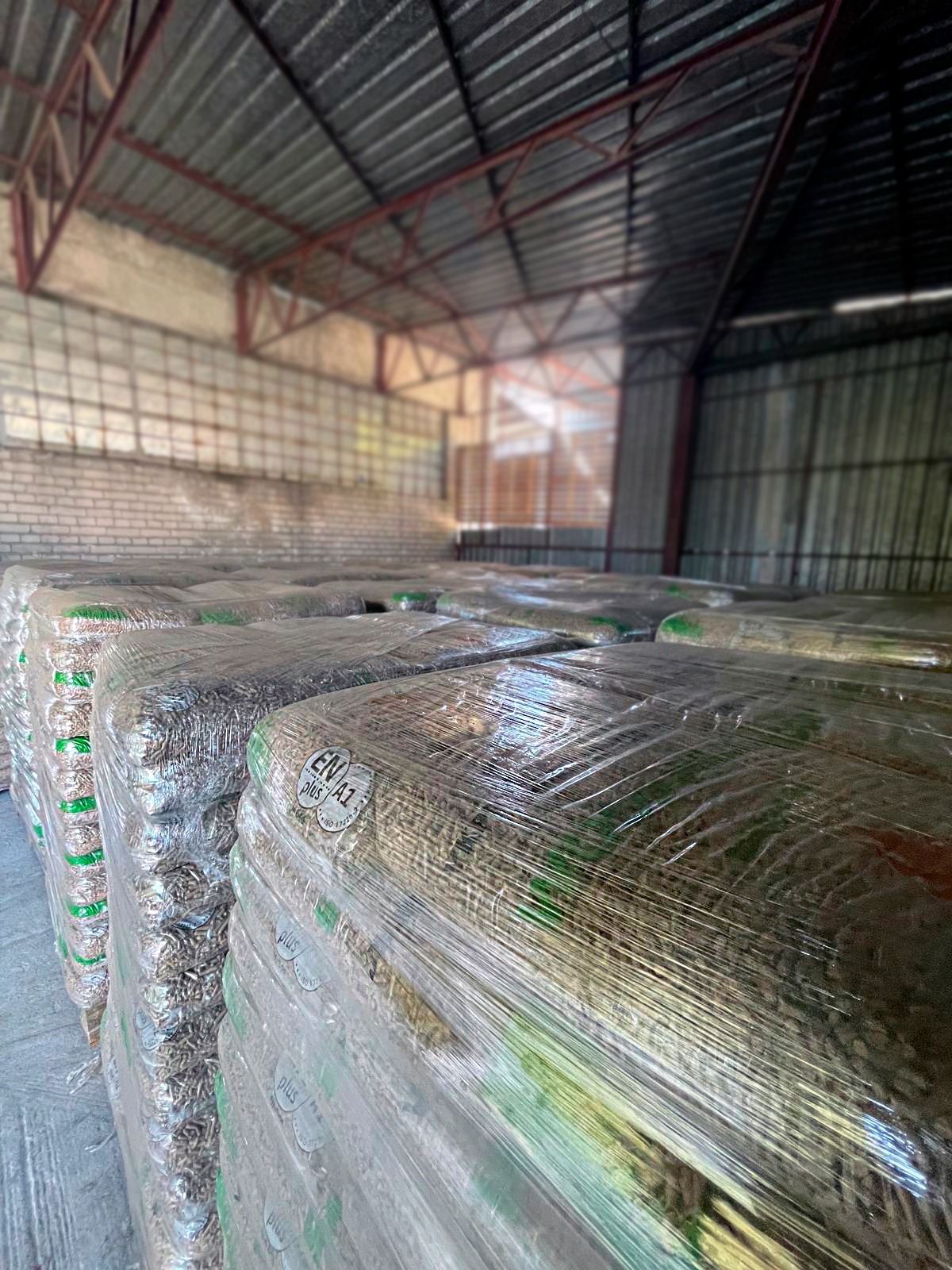 Skladowane, owinięte folią palety z pelletem drzewnym w magazynie z metalową konstrukcją dachu i ceglaną ścianą w tle. Na opakowaniach widoczne certyfikaty jakości.
