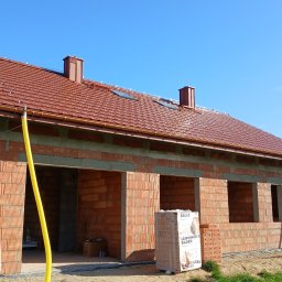 Marcin Łysakowski MARBUD - Niedokończony dom z czerwonej cegły z dachem z dachówki, widoczne okna i kominy, na pierwszym planie paleta cegieł. Żółty wąż na zewnątrz. Błękitne niebo w tle.