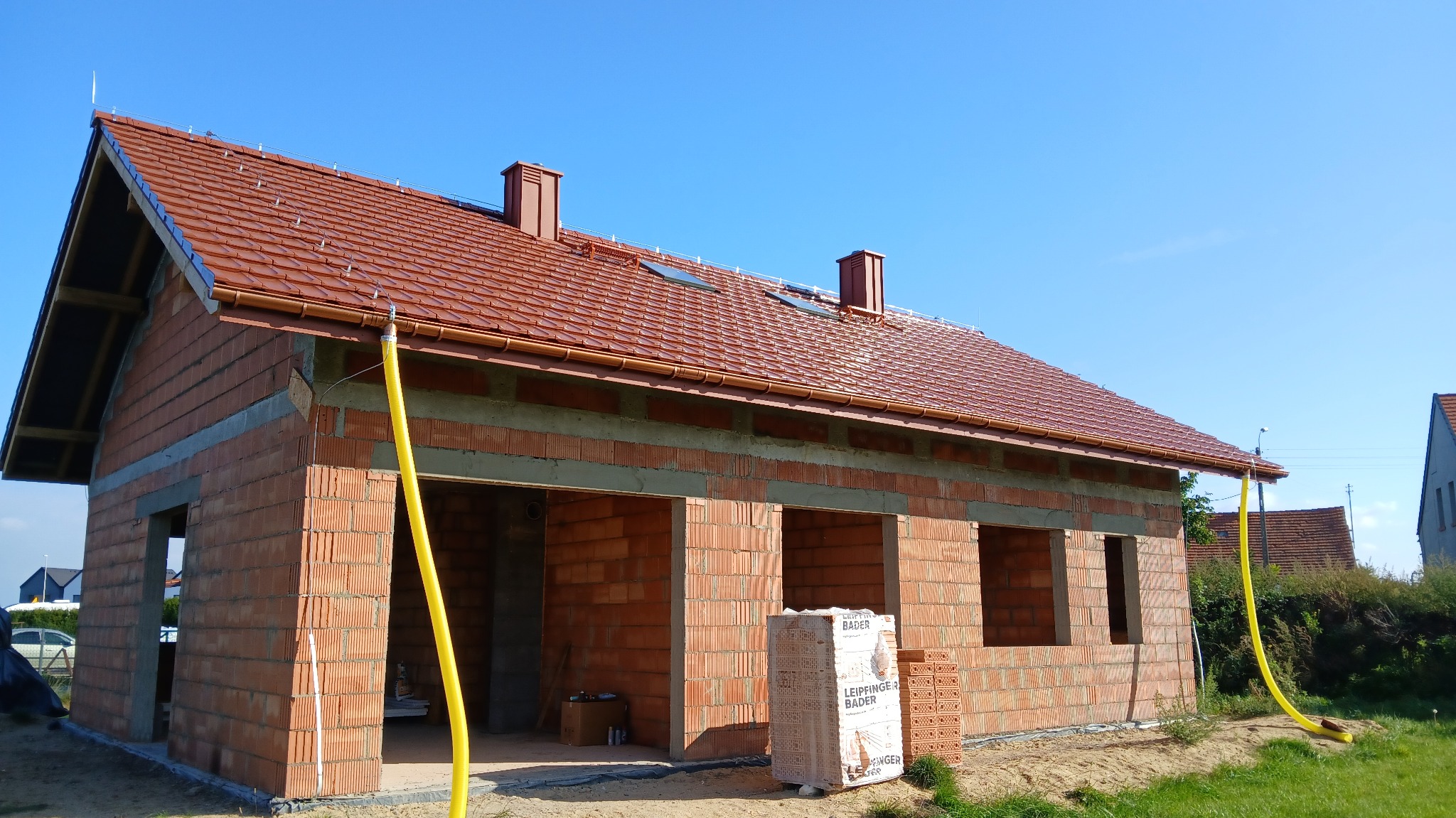 Niedokończony dom z czerwonej cegły z dachem z dachówki, widoczne okna i kominy, na pierwszym planie paleta cegieł. Żółty wąż na zewnątrz. Błękitne niebo w tle.