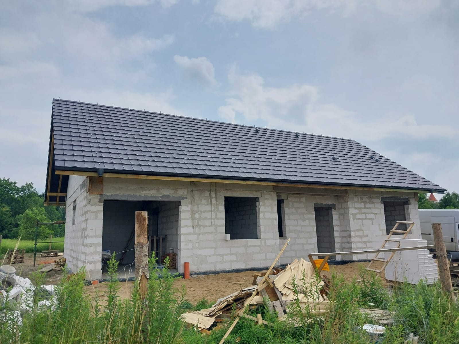 Surowy stan domu z białego pustaka z ciemnym dachem. Widoczne otwory okienne i drzwiowe, drewniane elementy konstrukcyjne. Na pierwszym planie gruz i roślinność.