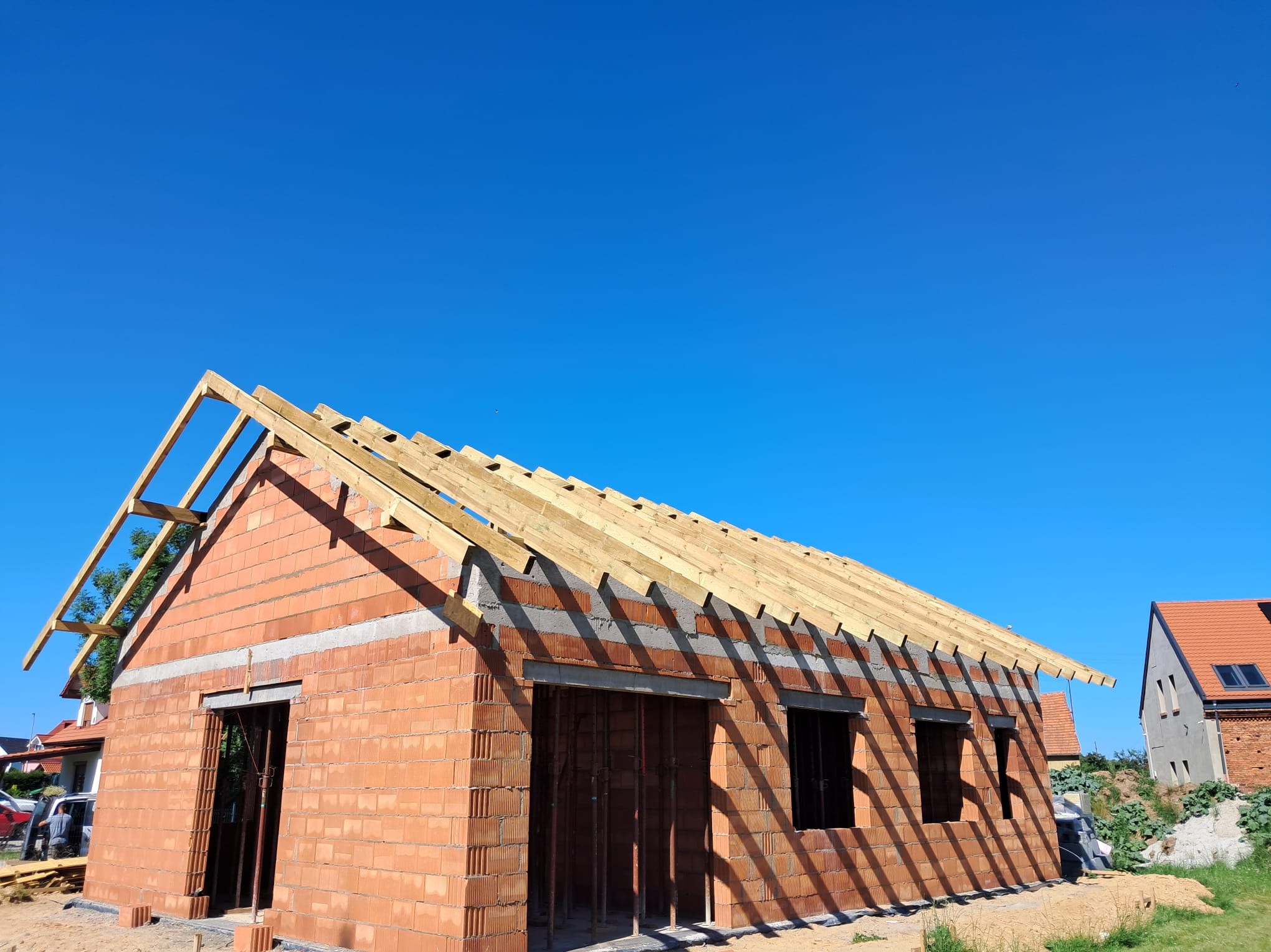 Budowa domu z czerwonej cegły w Wołowie, z widoczną konstrukcją dachu na tle błękitnego nieba. Etap murowania ścian i przygotowania do dalszych prac.
