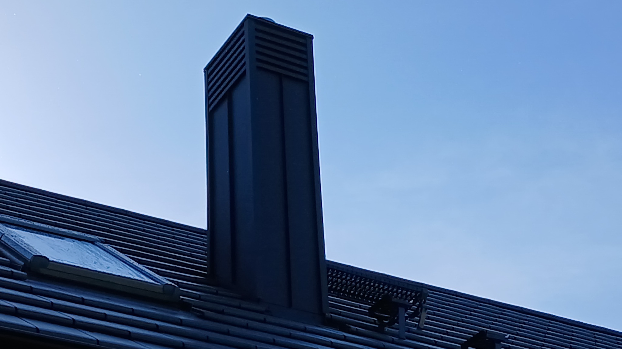Czarny komin wentylacyjny na dachu z dachówki ceramicznej, widok z dołu na tle błękitnego nieba. Widoczne okno dachowe i elementy systemu odwadniającego.