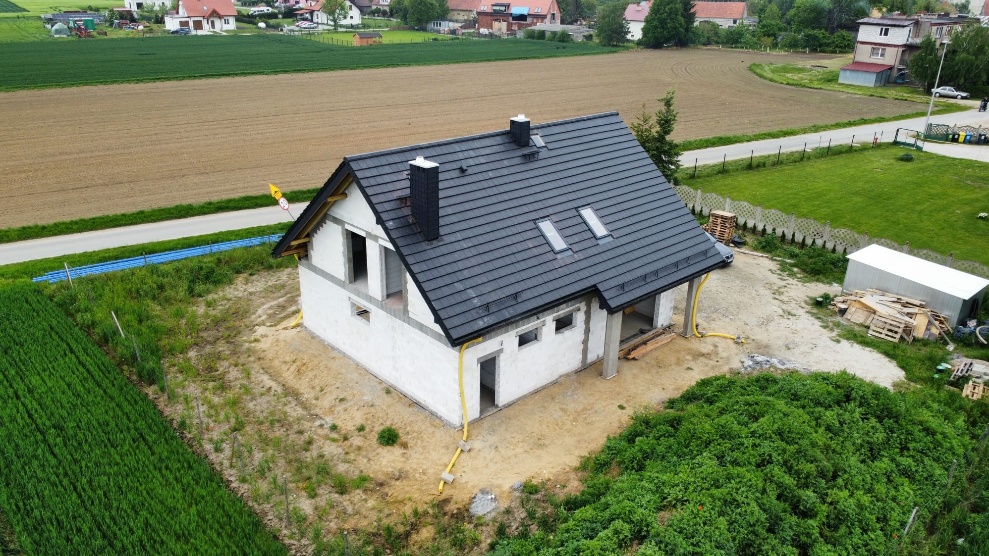 Dom w trakcie budowy z ciemną dachówką, widok z góry. Żółte rury przy ścianach, w tle pola i zabudowania. Stan surowy otwarty, brak okien.