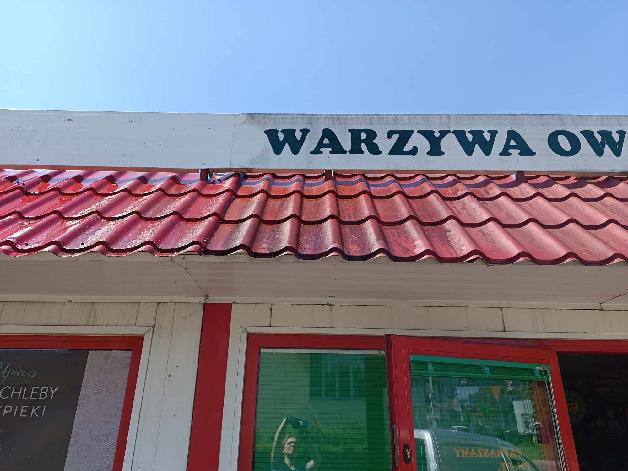 Czerwony dach pawilonu handlowego z napisem 'WARZYWA OWOCE' nad wejściem. Na oknach naklejki z wypiekami.