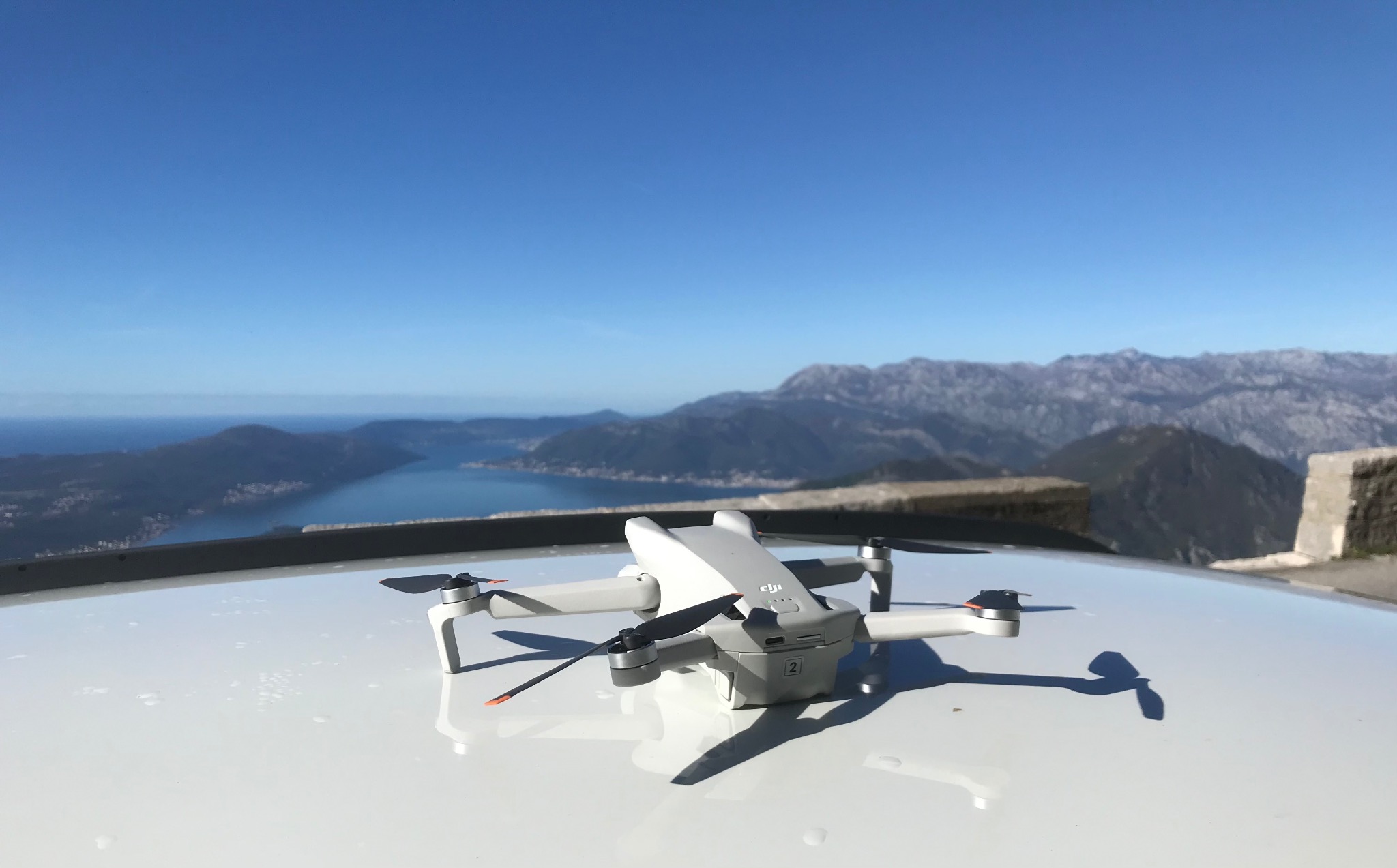 Biały dron DJI Mini 2 leży na białej powierzchni z widokiem na góry i zatokę w oddali, błękitne niebo w tle.