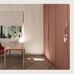 Józefina Furmańczyk - Wnętrze pokoju z biurkiem z jasnego drewna, lampką, książkami, roślinami w doniczkach na parapecie i dużą, różową szafą z ozdobnymi uchwytami.