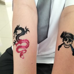 Bajkowe Animacje dla Dzieci Małopolska - Dziecięce ramiona z brokatowymi tatuażami: czarno-różowy smok i piracka czaszka z skrzyżowanymi piszczelami w kolorze niebieskim. Atrakcja dla dzieci na imprezie.