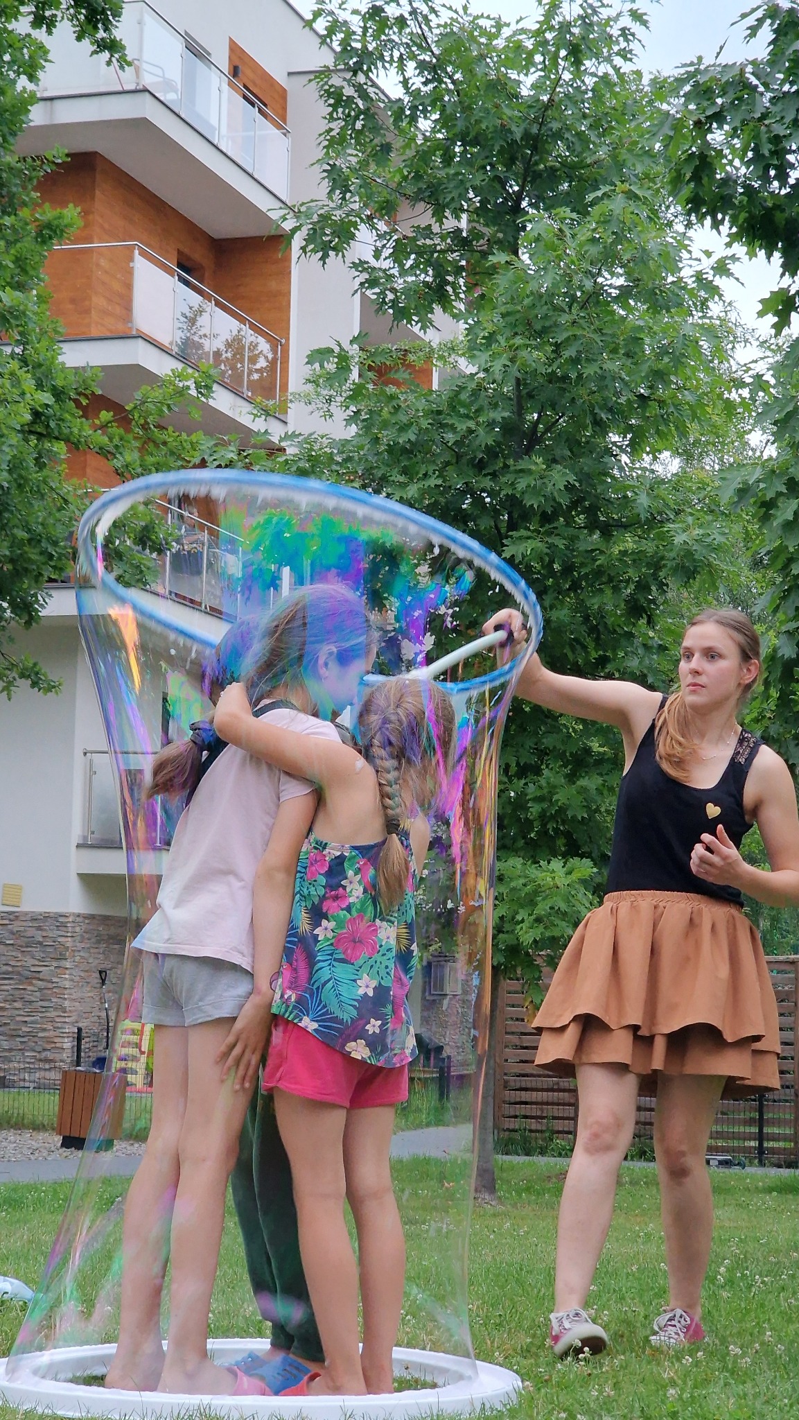 Animator tworzy ogromną bańkę mydlaną, w której zamknięte są dwie dziewczynki. Kobieta w brązowej spódnicy trzyma kij do robienia baniek. Zabawa na trawie.