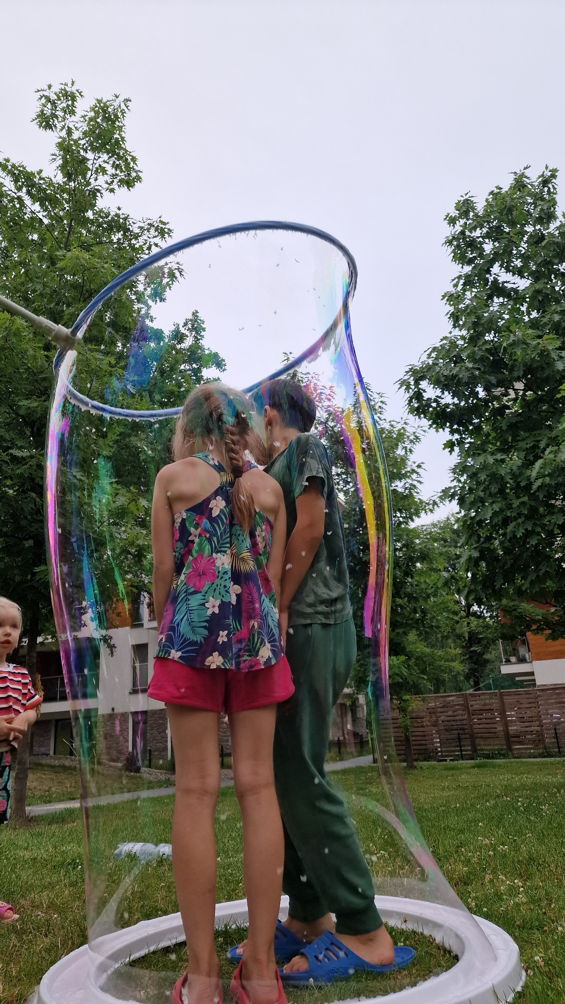 Dzieci wewnątrz gigantycznej bańki mydlanej, tęczowe refleksy na powierzchni, zielony trawnik i drzewa w tle, zabawa na świeżym powietrzu.