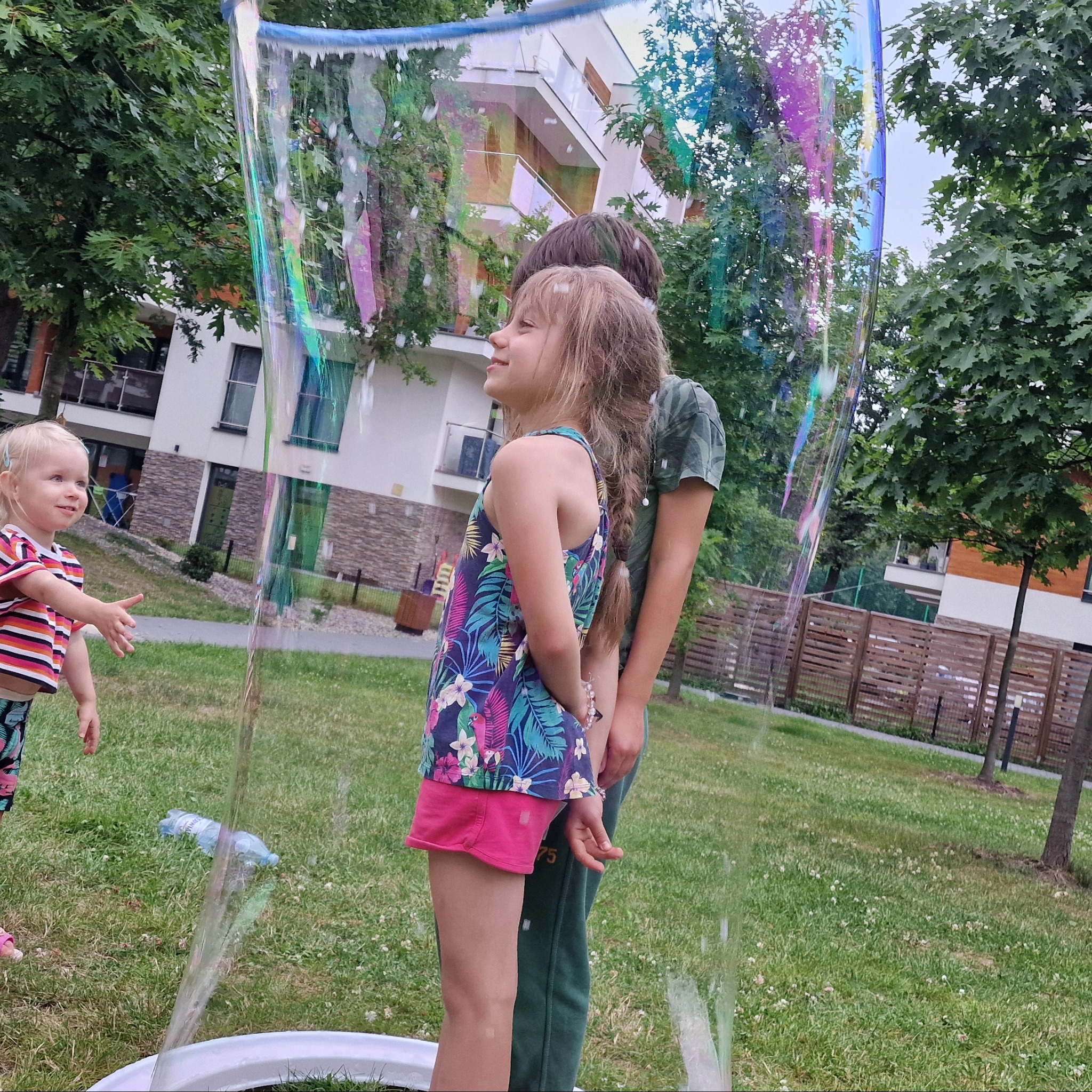 Dzieci w gigantycznej bańce mydlanej na trawniku przed budynkiem. Dziewczynka w różowych spodenkach i kwiecistym topie patrzy w górę, obok stoi chłopiec. Mała dziewczynka wyciąga ręce.