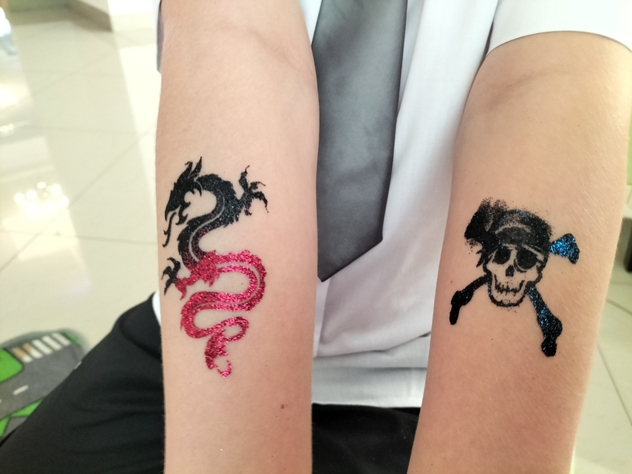 Dziecięce ramiona z brokatowymi tatuażami: czarno-różowy smok i piracka czaszka z skrzyżowanymi piszczelami w kolorze niebieskim. Atrakcja dla dzieci na imprezie.