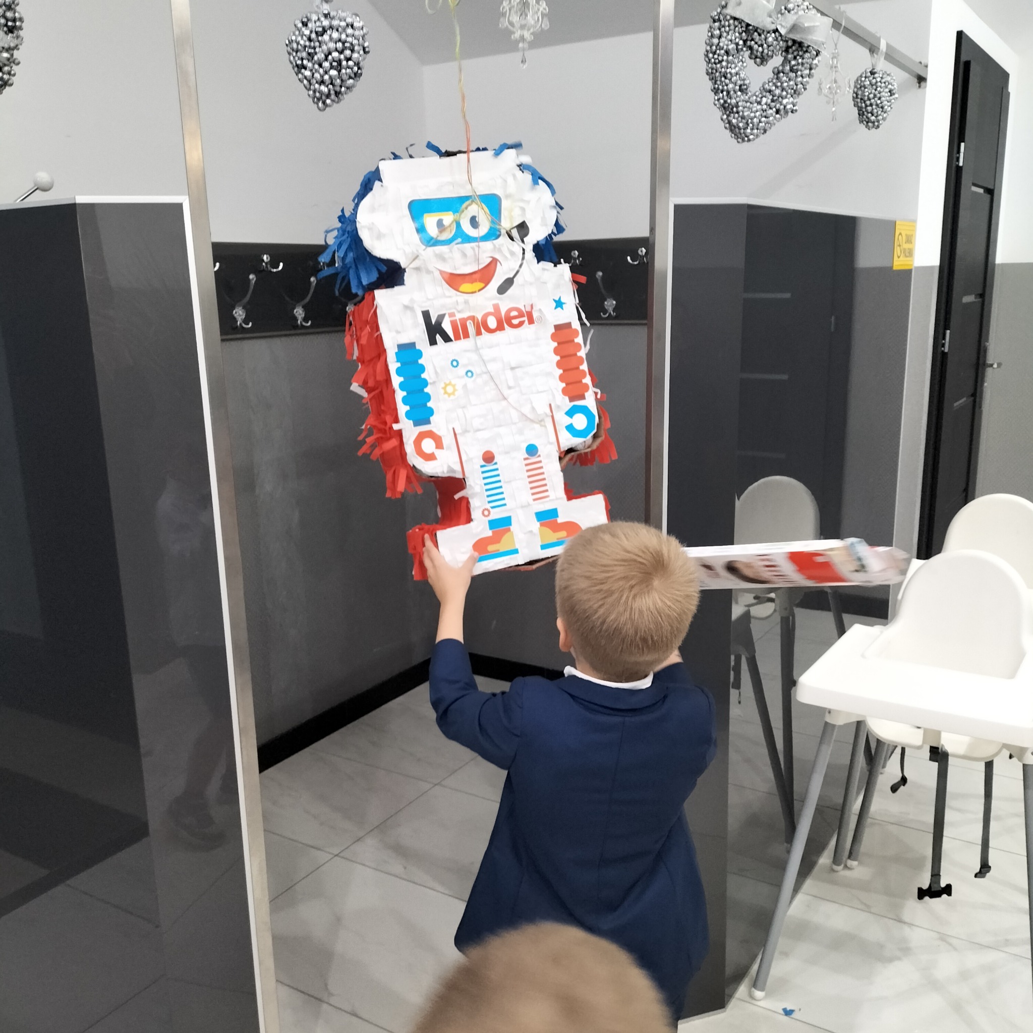 Chłopiec w marynarce podnosi piñatę w kształcie robota Kinder na tle sali z dekoracjami. Przygotowania do zabawy urodzinowej.