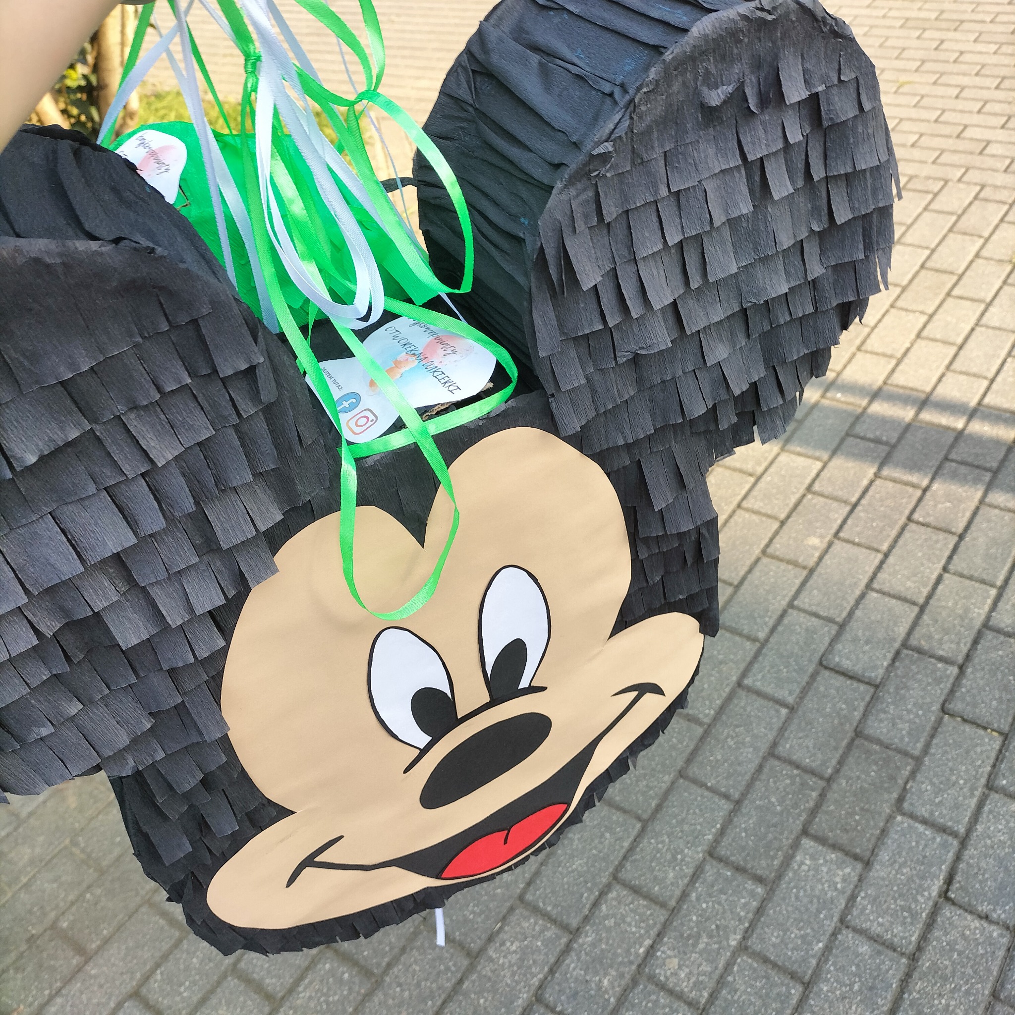 Pinata w kształcie Myszki Miki z czarnymi frędzlami i kolorowymi wstążkami, gotowa do zabawy na imprezie. Widoczne logo firmy na etykiecie.