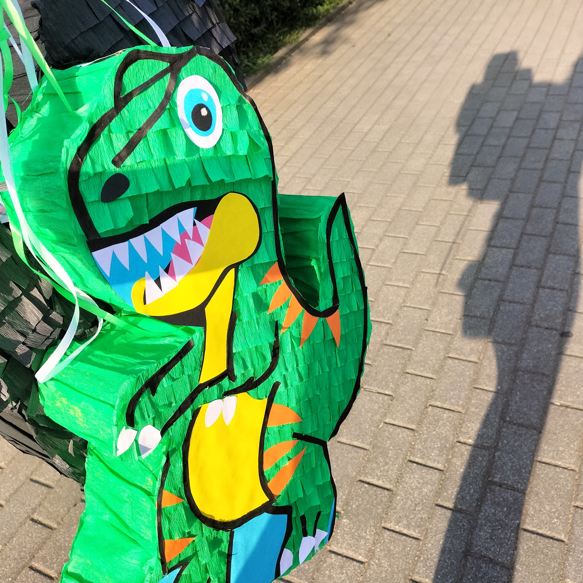 Zielona piniata w kształcie dinozaura z kolorowymi detalami, stojąca na chodniku, rzucająca cień. Dekoracja urodzinowa dla dzieci, impreza plenerowa.