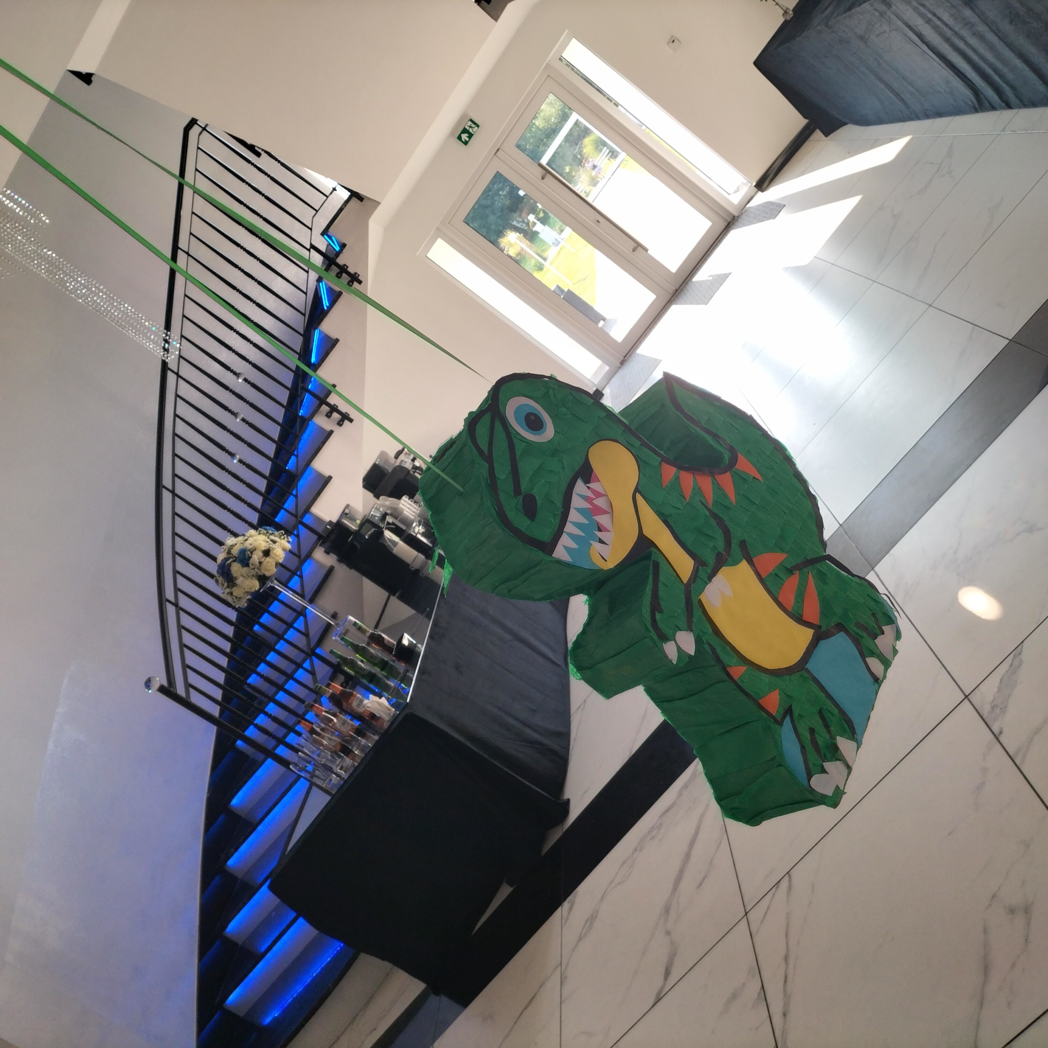 Zielona pinata w kształcie dinozaura zawieszona w eleganckim wnętrzu z marmurową podłogą i schodami z niebieskim podświetleniem, przygotowana na imprezę.